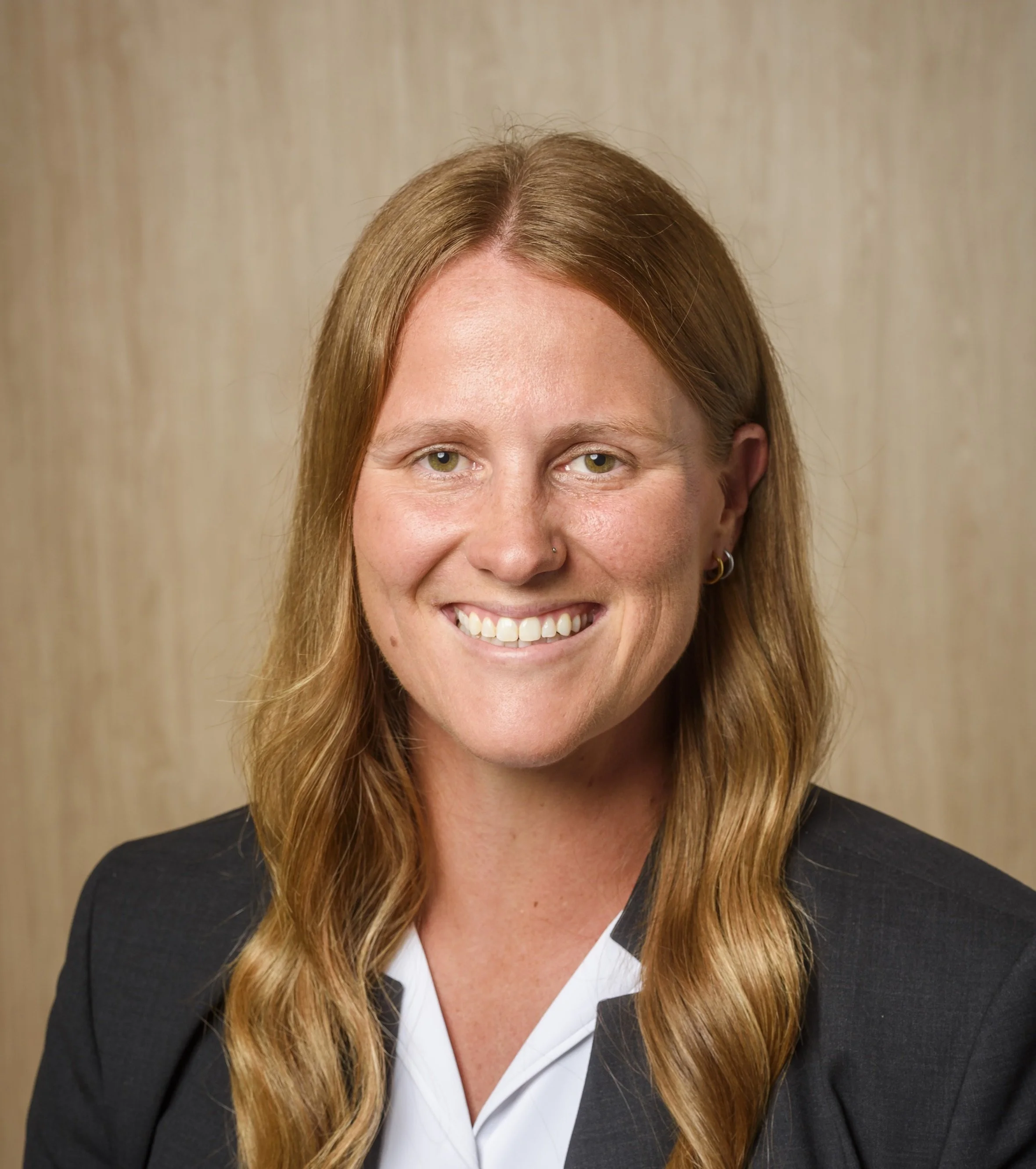 A woman with long red hair smiling, wearing earrings and a black blazer, against a beige background.