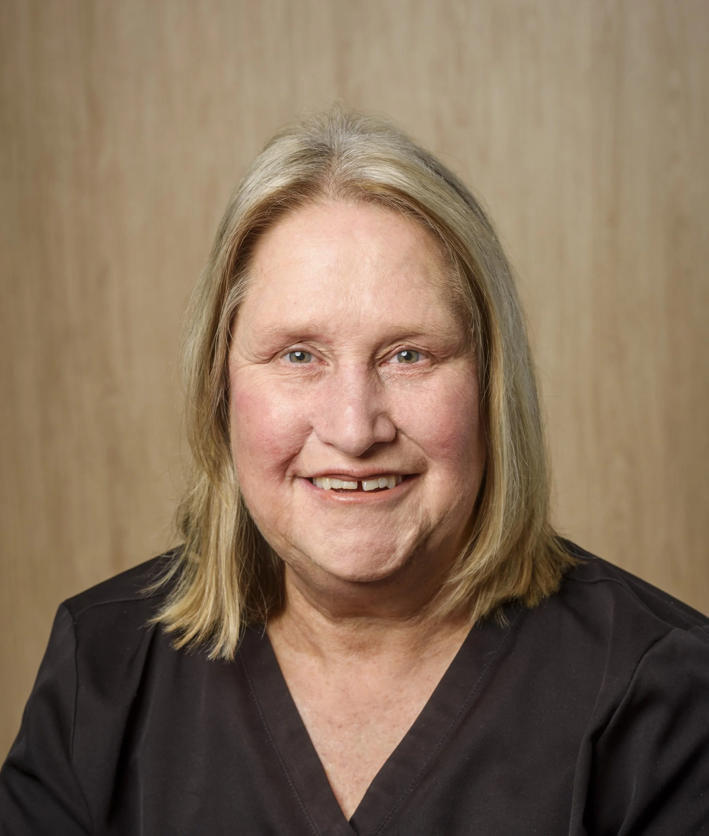 Close-up portrait of a middle-aged woman with shoulder-length light brown hair, wearing a black jacket and white shirt, smiling at camera against a neutral background.