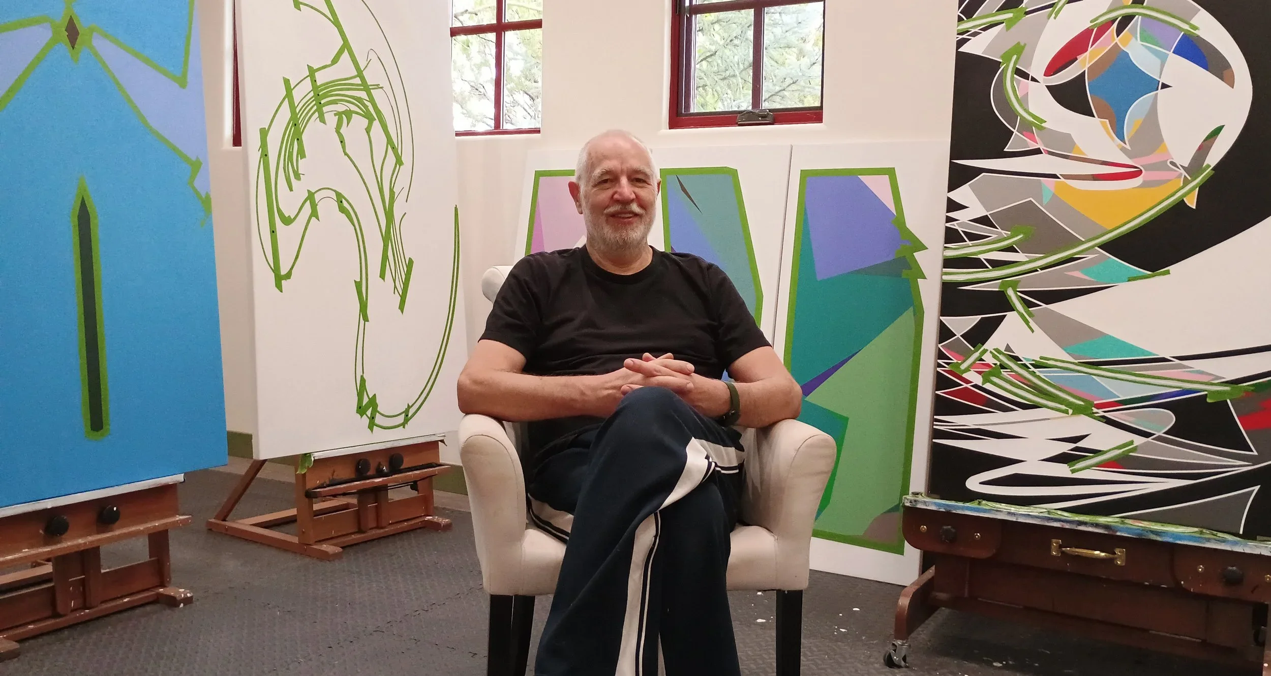 An older man with a beard, wearing a black t-shirt, sitting with his legs crossed in a white armchair in an art studio. Behind him are multiple large abstract paintings with vibrant colors and bold designs, on easels and leaning against the wall.