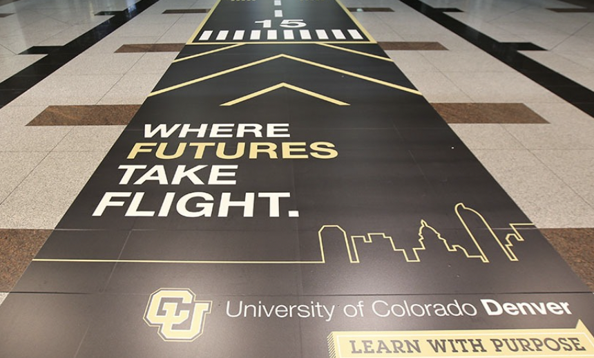 A black and yellow advertisement floor decal at Denver International Airport promoting the University of Colorado Denver with the slogan "Where futures take flight," featuring a city skyline outline and the university's logo.