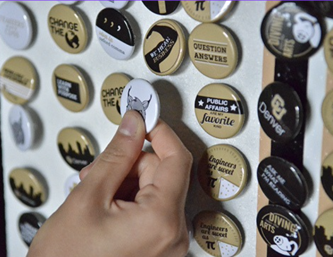 Person holding a button with a cartoon character. Background of various buttons on a wall, with sayings and designs including 'Change the', 'Question Answers', 'Public Affairs favorite', and 'Engineers are sixed.'