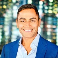 A man with dark hair in a blue suit smiling in front of a cityscape background.