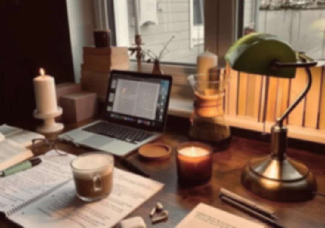 A cozy workspace with a wooden desk near a window, featuring a laptop, a lit candle, a cup of coffee, books, a desk lamp, and various office supplies.