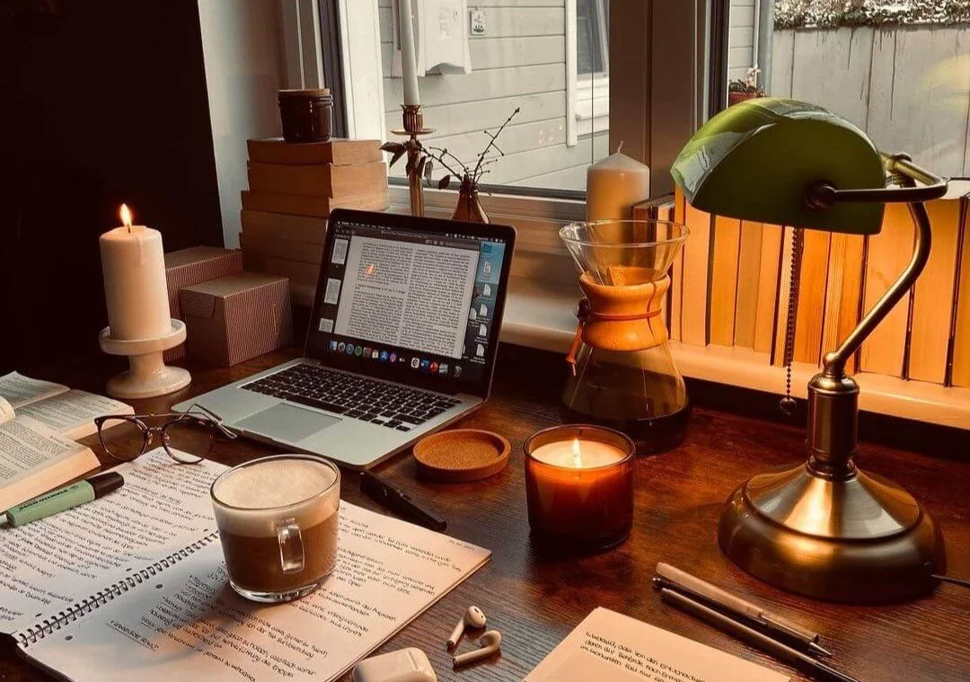 A cozy workspace with an open laptop, books, a cup of coffee, glasses, candles, a desk lamp, and a window with a view outside.