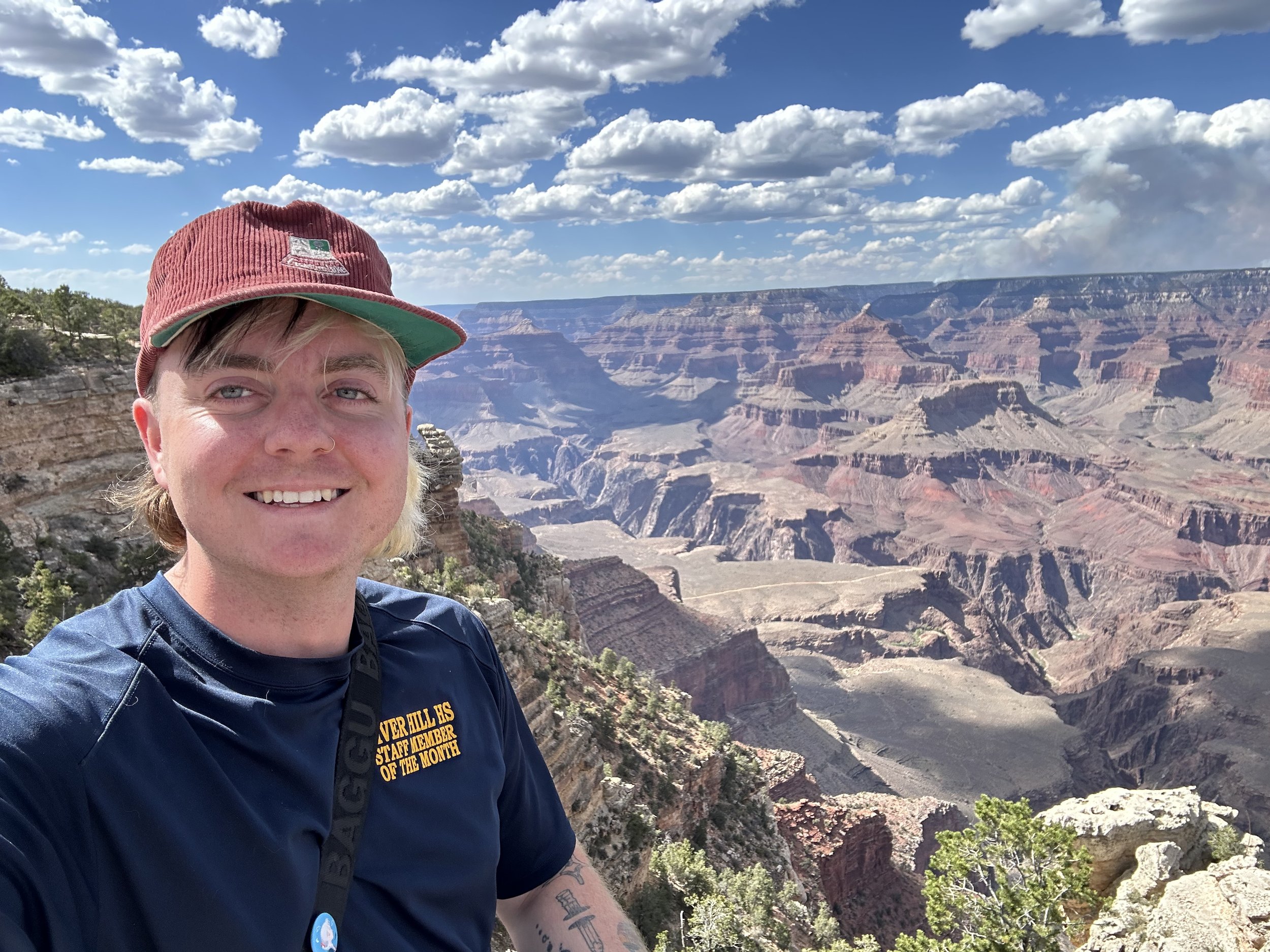 Jade E Welsh, wearing a burgundy cap and a blue shirt, standing at the Grand Canyon, with the canyon's layered rock formations and a partly cloudy sky in the background.