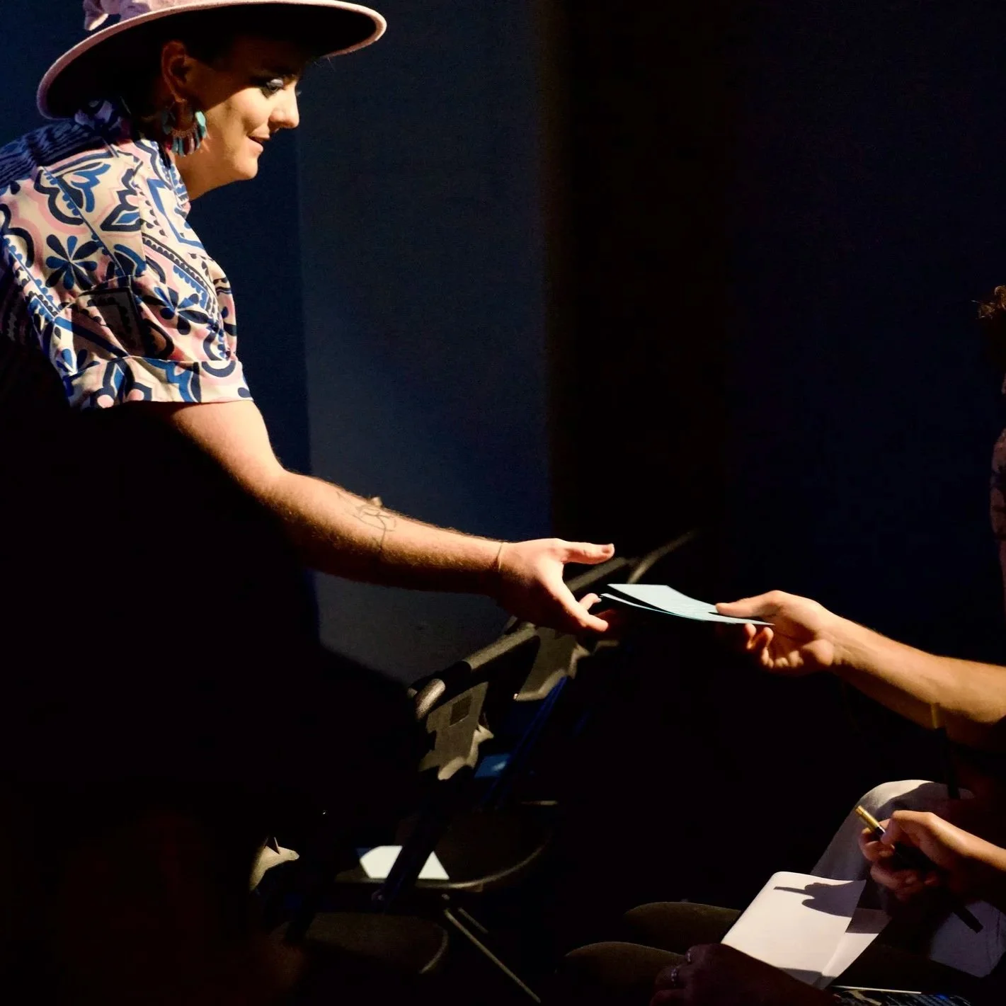 Jade Ethan wearing a hat and a patterned shirt is handing a document or paper to another person in a dark setting.