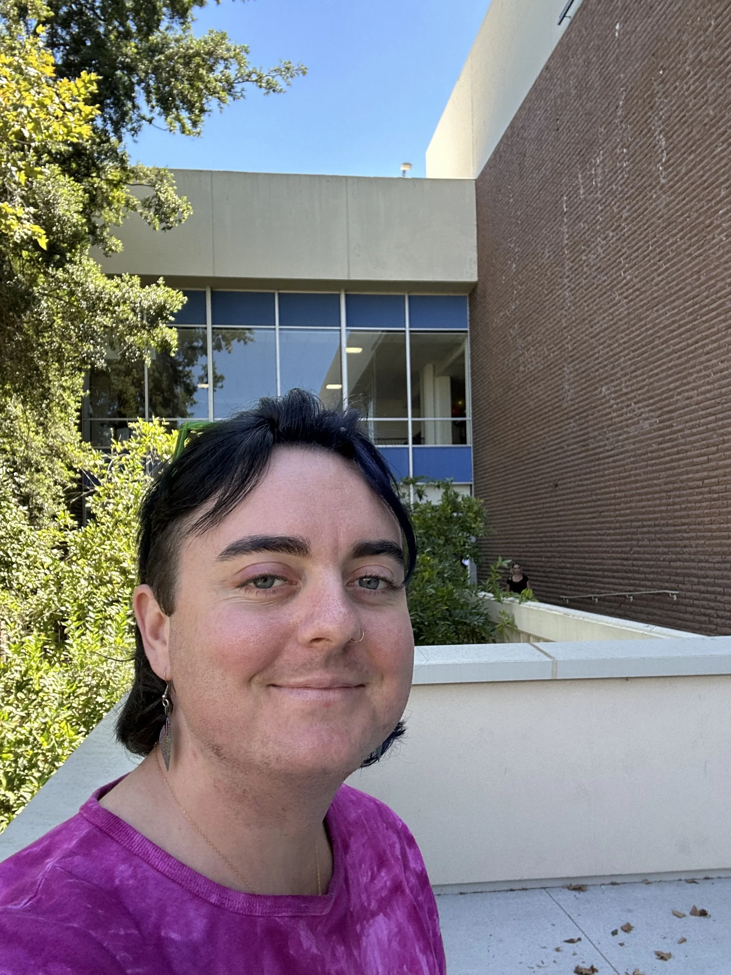 A photo of Jade E Welsh, who has dark hair and is wearing a purple shirt, standing outside a modern building with large windows and brick walls, surrounded by greenery, on a sunny day.