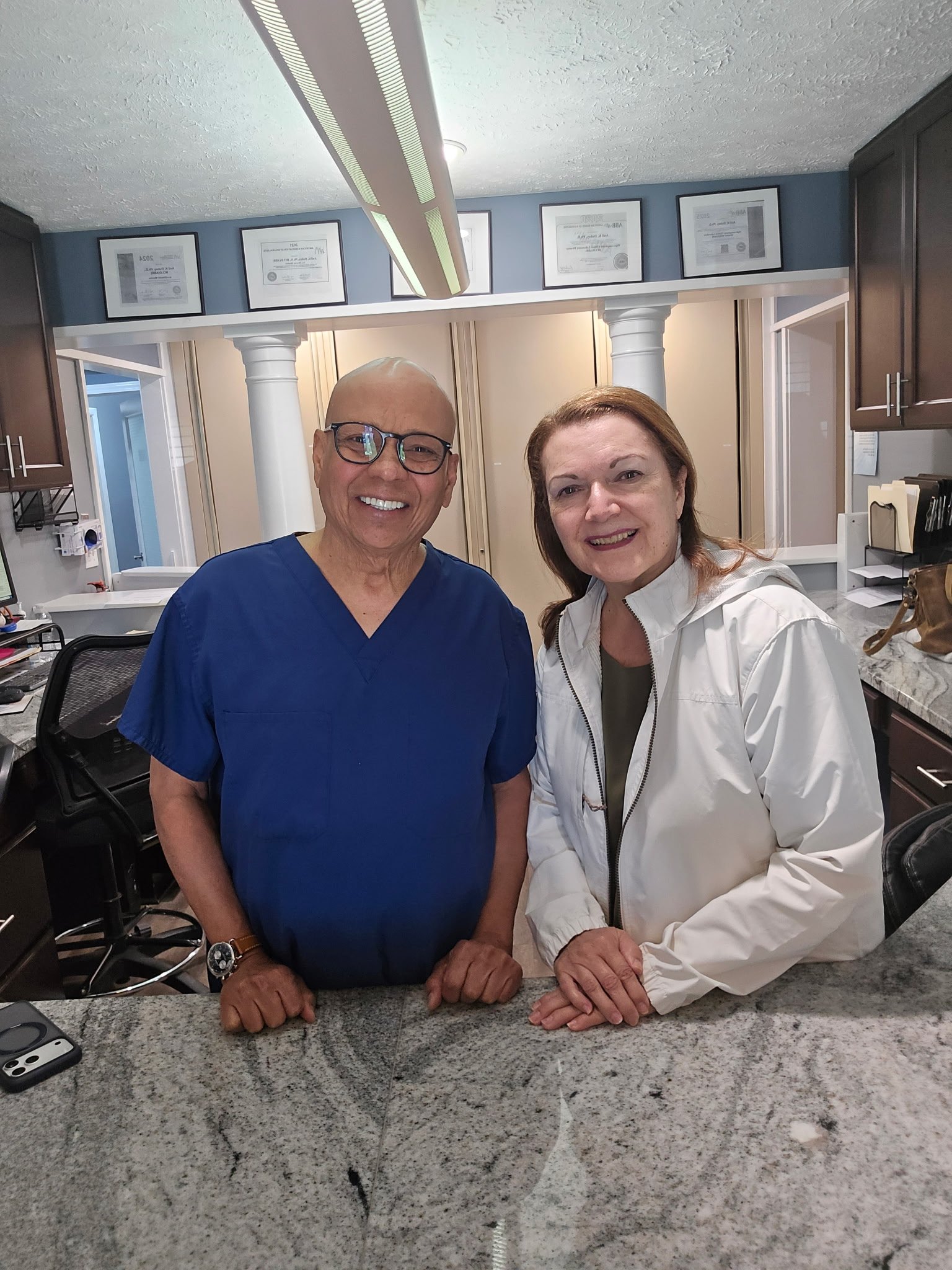 A man wearing glasses and a blue medical scrubs and a woman with auburn hair and a white jacket are smiling and posing for a selfie in a medical or office reception area. They are standing behind a marble counter, with framed certificates on the wall behind them and a modern light fixture hanging from the ceiling.