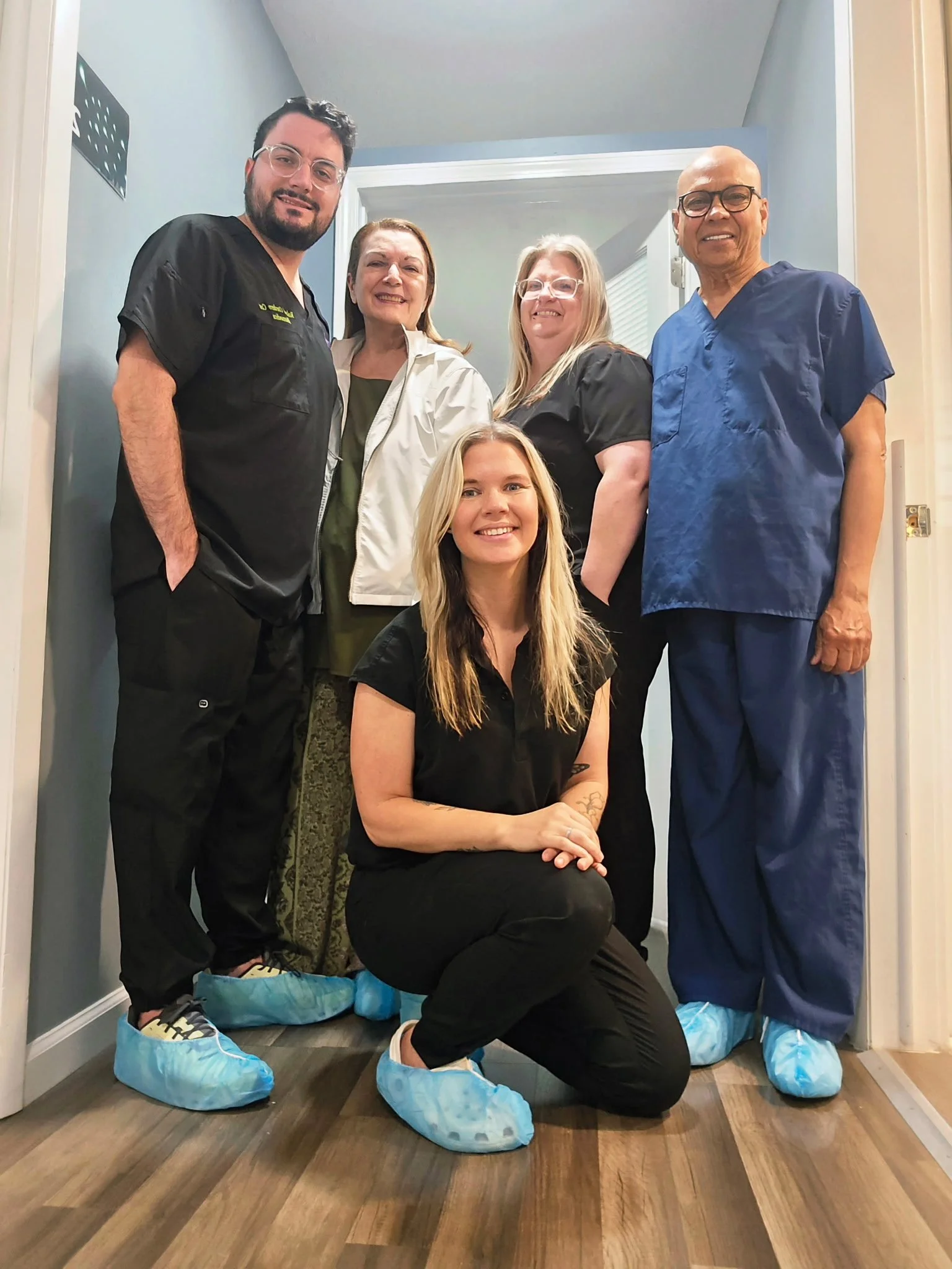 Group of five people, including medical professionals, standing in a hallway, wearing blue shoe covers, with one person kneeling in front. They are smiling and posing for a photo.