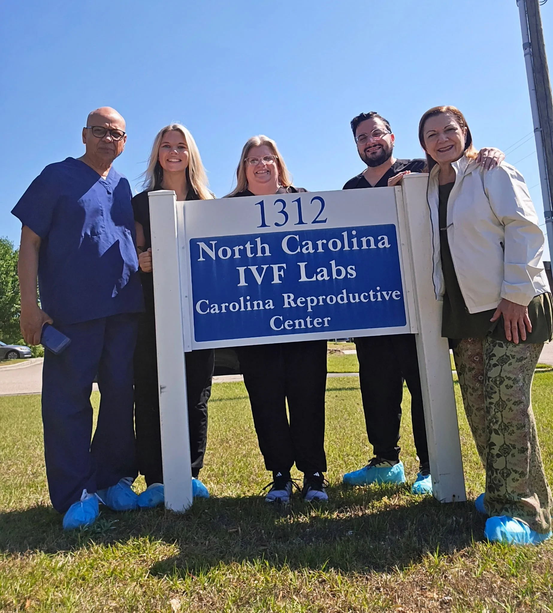 Group of five people standing outdoors behind a sign that reads '1312 North Carolina IVF Labs' with a clear blue sky overhead. They are dressed in casual clothing and wearing blue shoe covers, smiling for the photo.