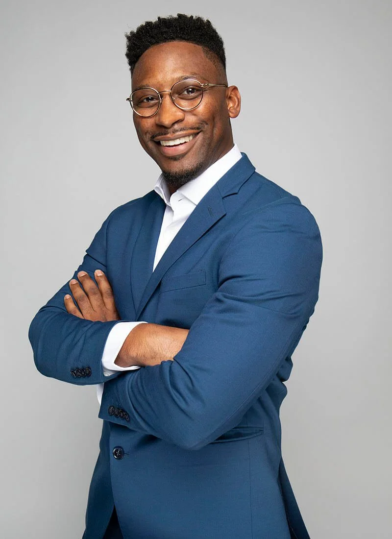 Smiling man in a blue suit and glasses with arms crossed against a gray background.