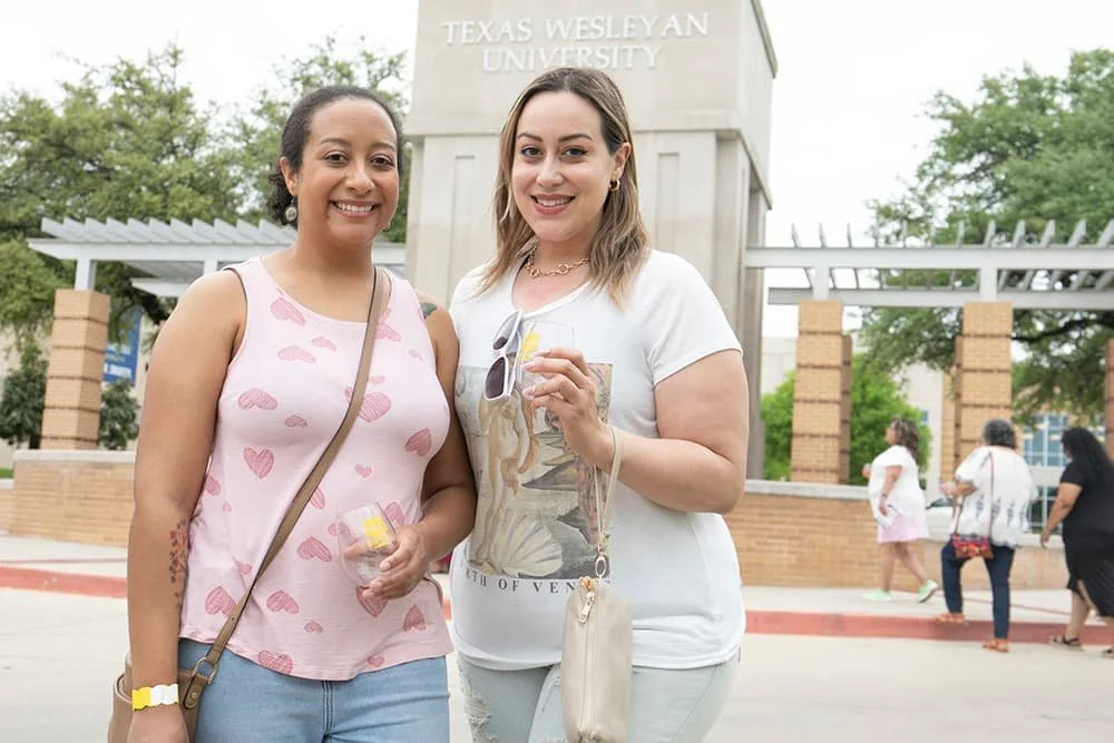 Two women holding drinks and smiling in front of Texas Wesleyan University sign
