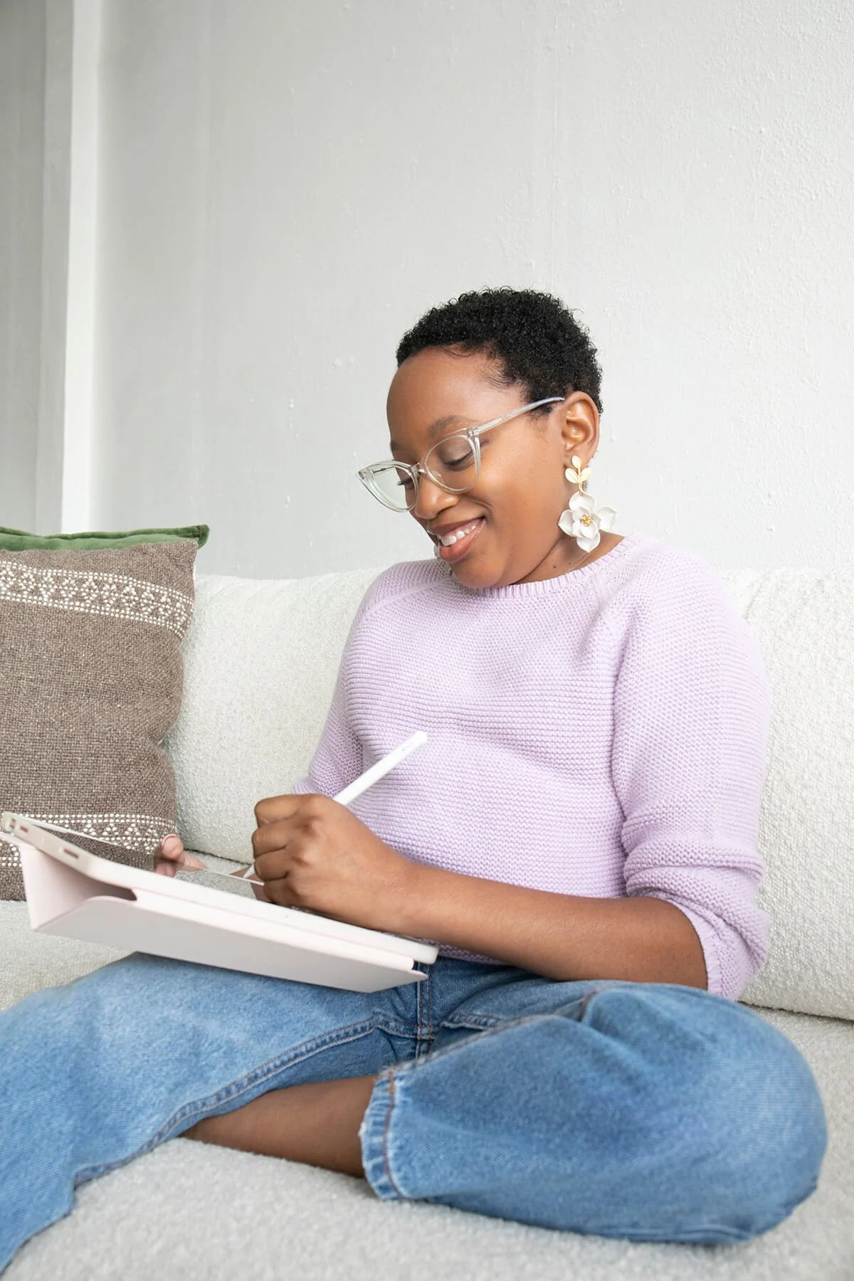 Person sitting on a couch writing on a tablet with a stylus, wearing a lilac sweater, jeans, glasses, and decorative earrings.