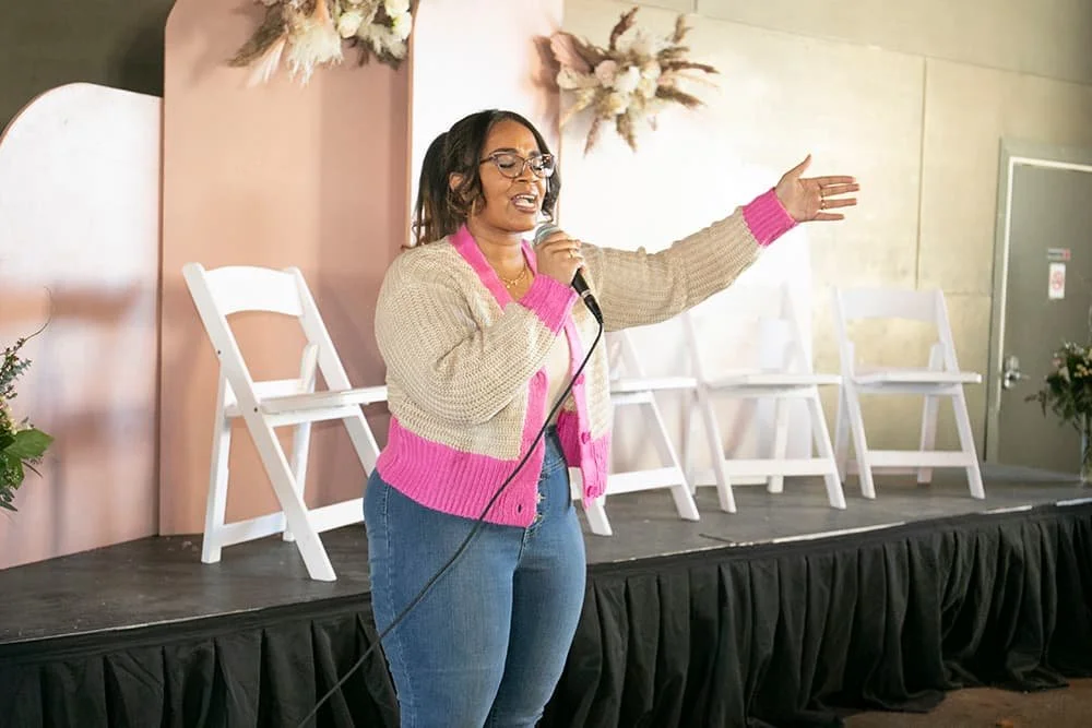 Woman speaking into a microphone on a stage with empty chairs and floral decorations.