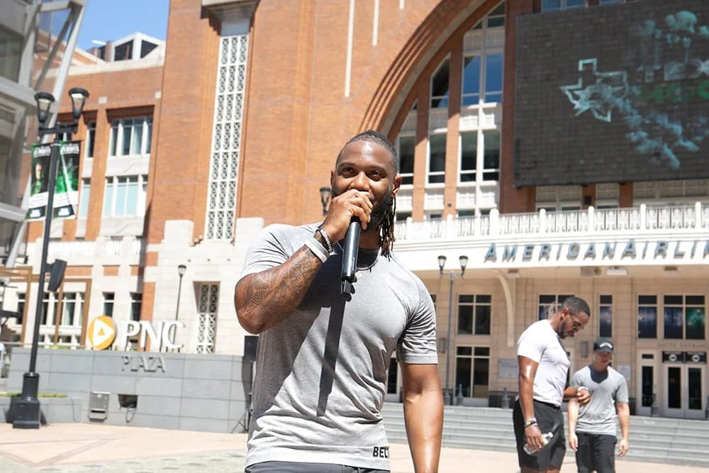 A man holding a microphone speaking outdoors near the American Airlines Center in Dallas, with people and buildings in the background.