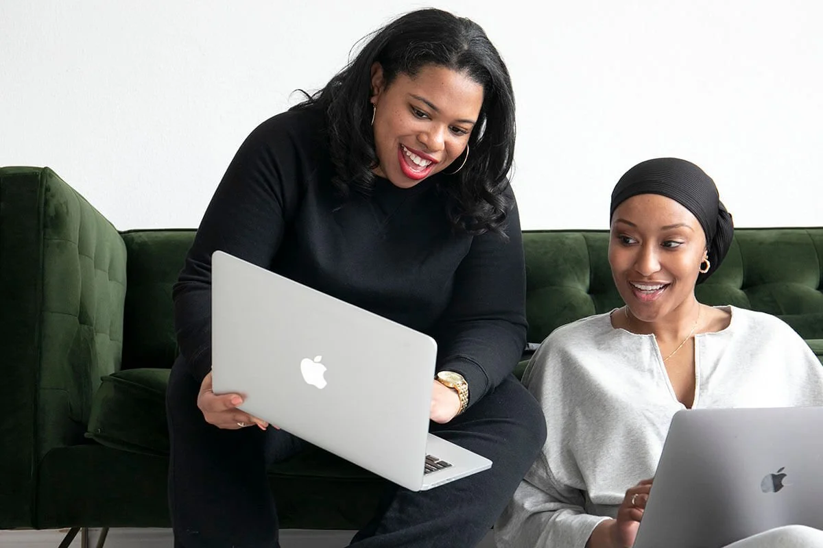 Two women smiling and looking at laptops, sitting on a green sofa.