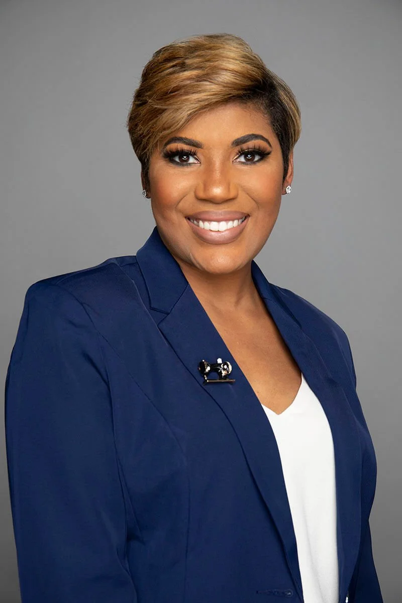 Portrait of a person with short hair wearing a navy blazer and white shirt, smiling against a gray background.