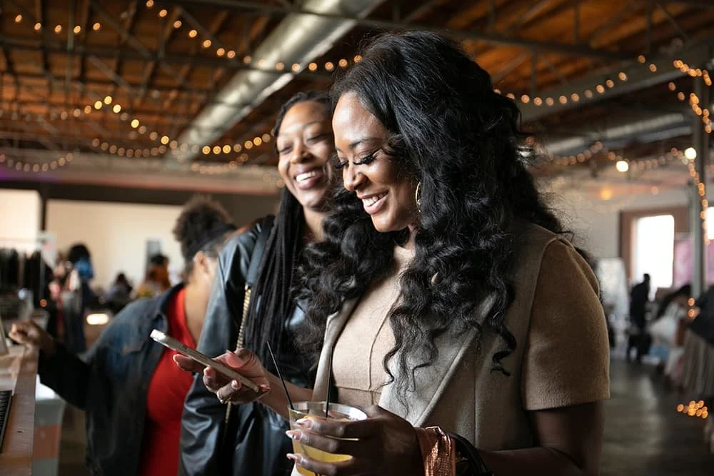 Two women smiling and looking at a smartphone in a warmly lit indoor space with string lights.