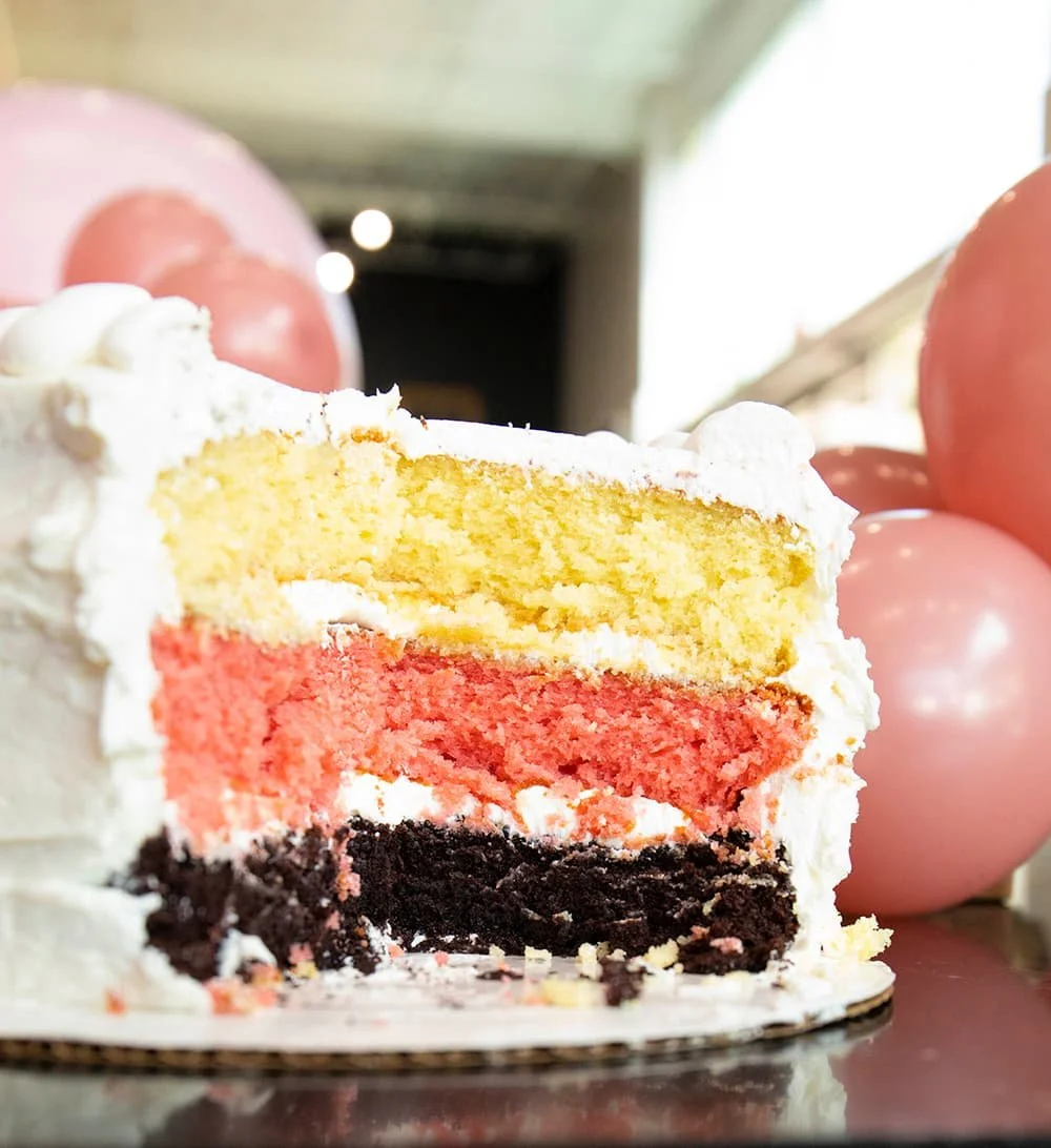 Three-layer cake with yellow, pink, and brown layers, topped with white icing, surrounded by pink balloons.