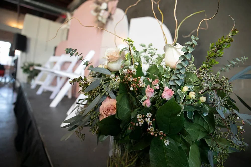 Floral arrangement with roses and greenery, white chairs in the background, indoor setting.