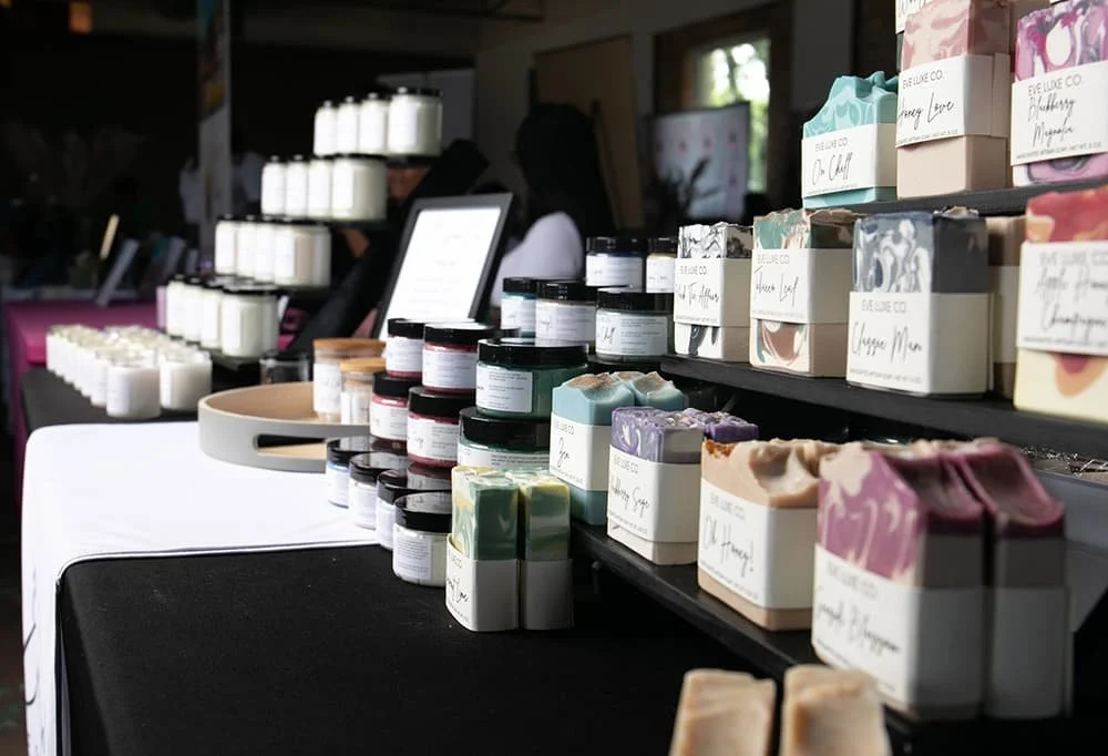 Display of handmade soaps, candles, and cosmetic jars on a table at a market stall.