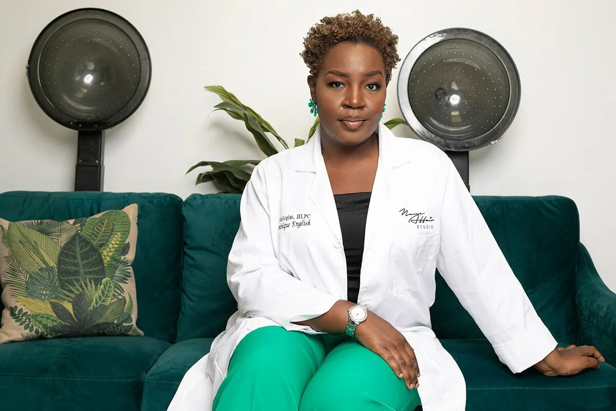 A person seated on a green sofa wearing a white lab coat, with hair styling equipment in the background, and a pillow with leaf designs.