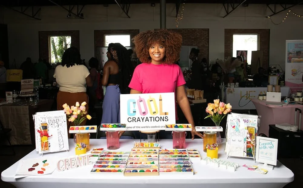 A woman stands behind a display table with colorful crayon products and a "Cool Crayations" sign at an indoor market or event. The table features various crayon shapes, small display stands, and decorative flowers. Other vendors and attendees are visible in the background.