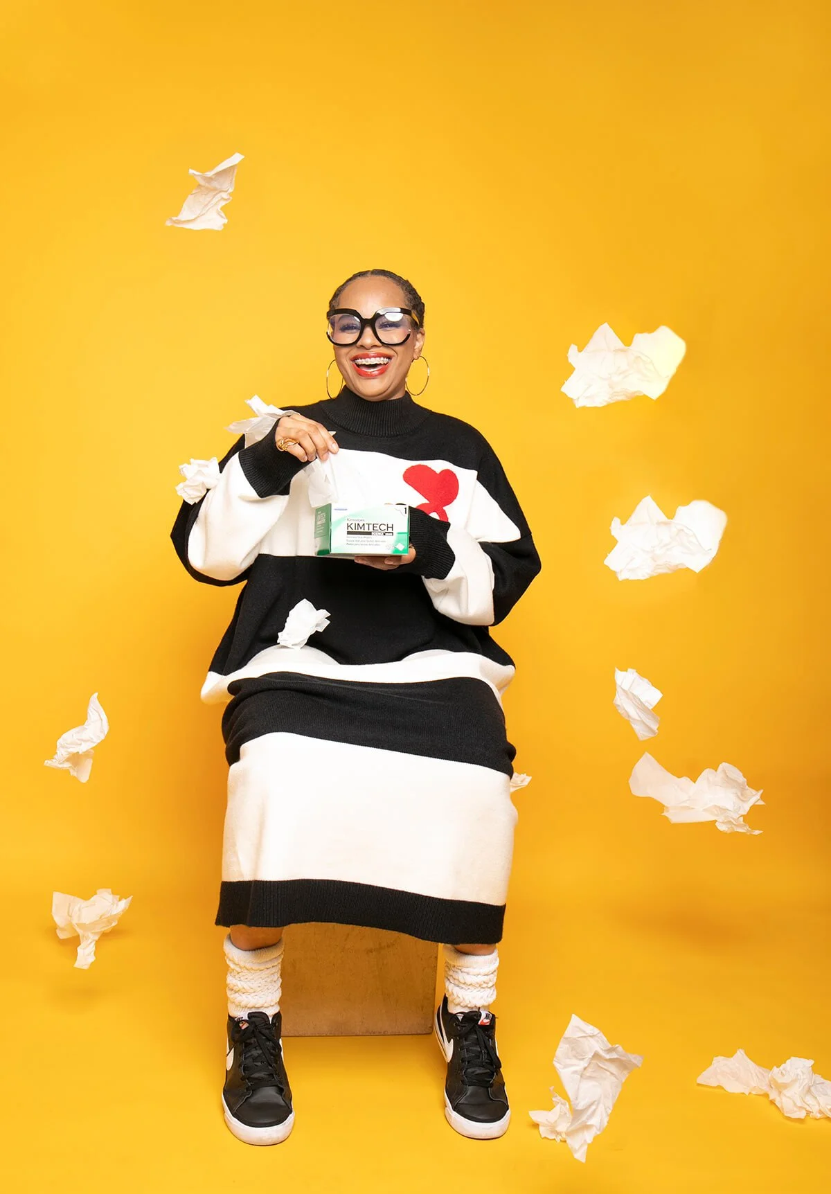 Person smiling and wearing a black and white striped sweater dress with a heart design, holding a box of Kimtech tissues, with tissues floating around against a yellow background.