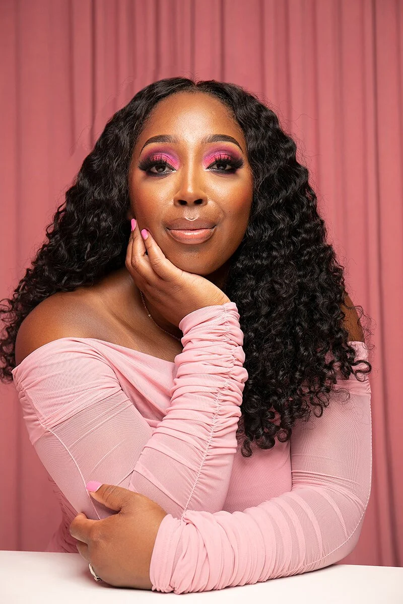 Woman in pink off-shoulder top with curly hair, resting chin on hand against a pink curtain background.