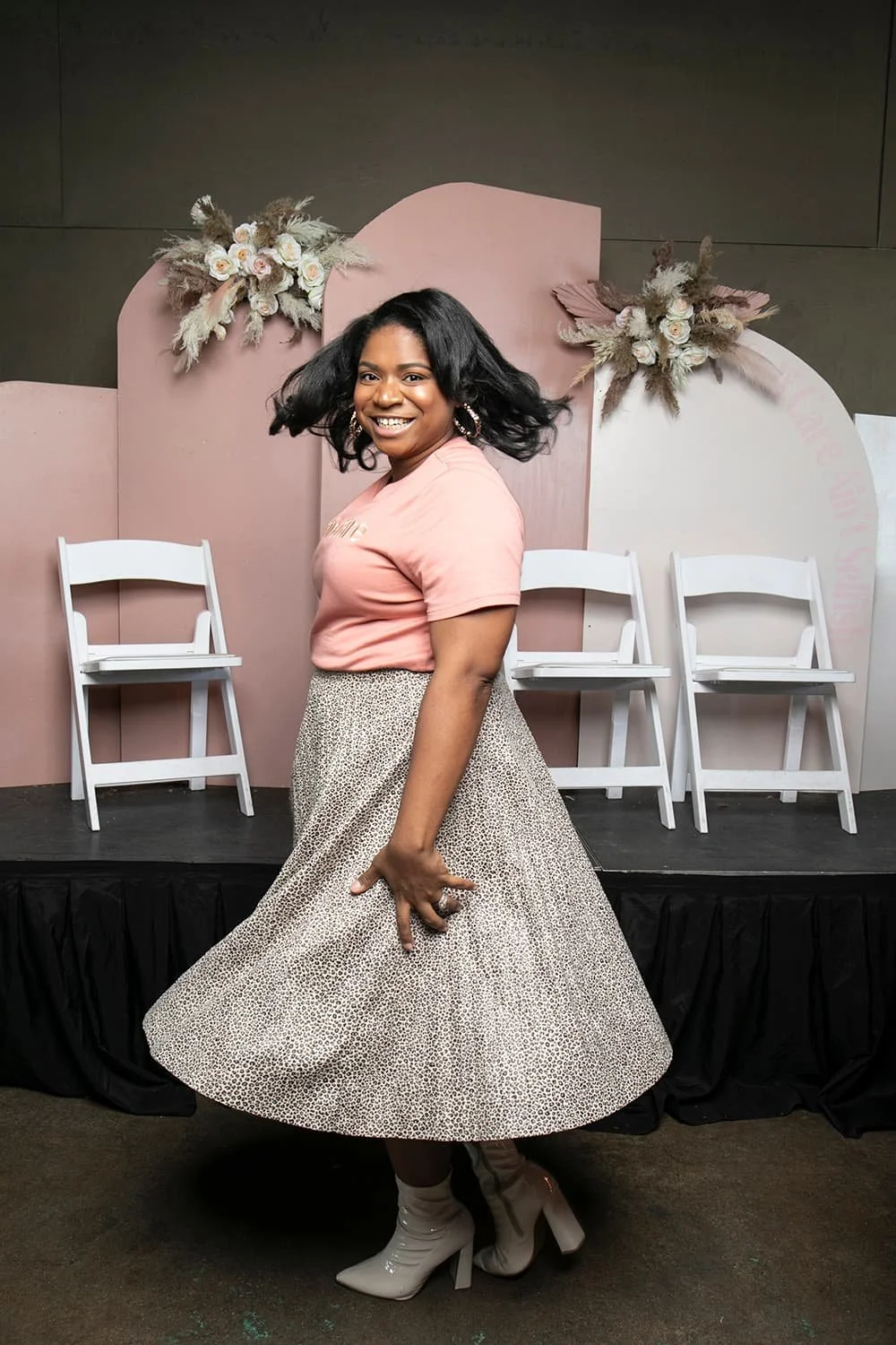 Woman smiling and twirling in a patterned skirt and pink top, in front of white chairs and decorative flowers on a stage.