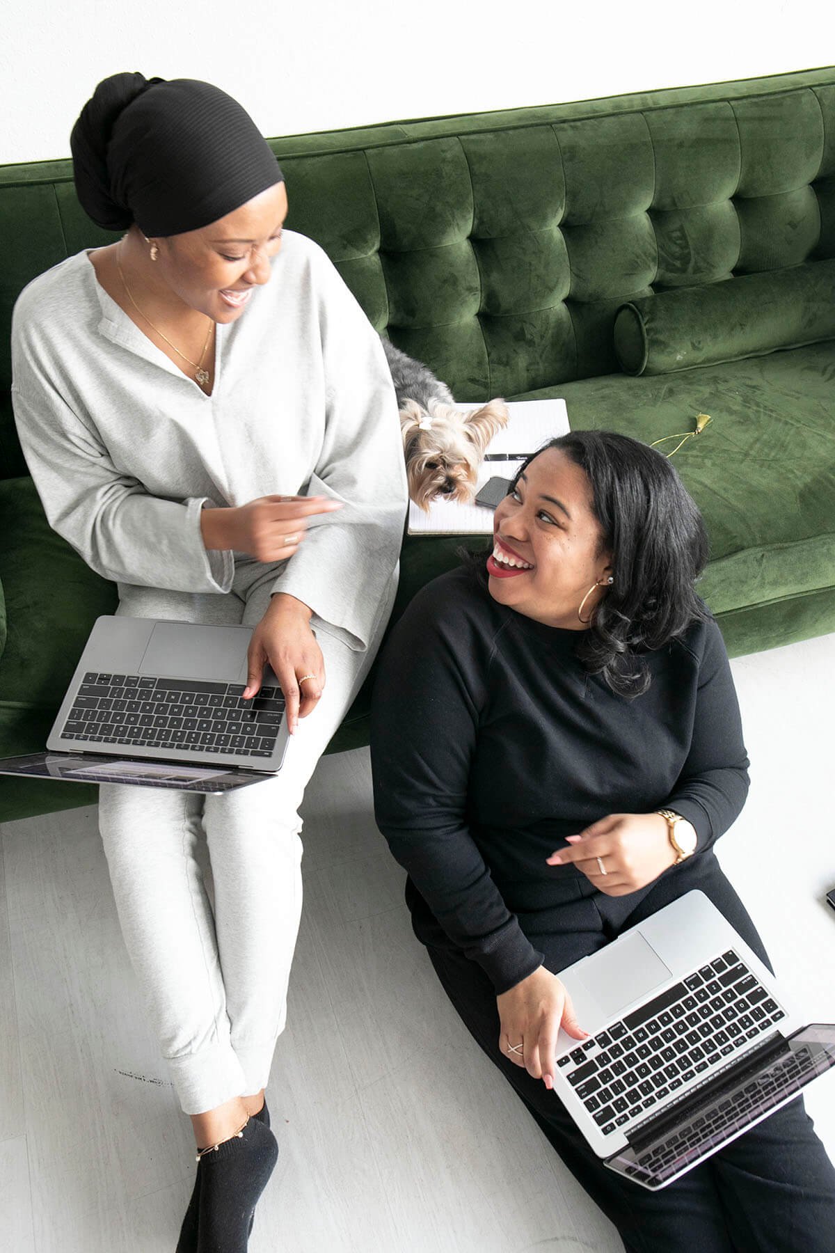 Two people sitting together on the floor with laptops, smiling and engaging in conversation. A small dog is on the sofa behind them with a notebook nearby.