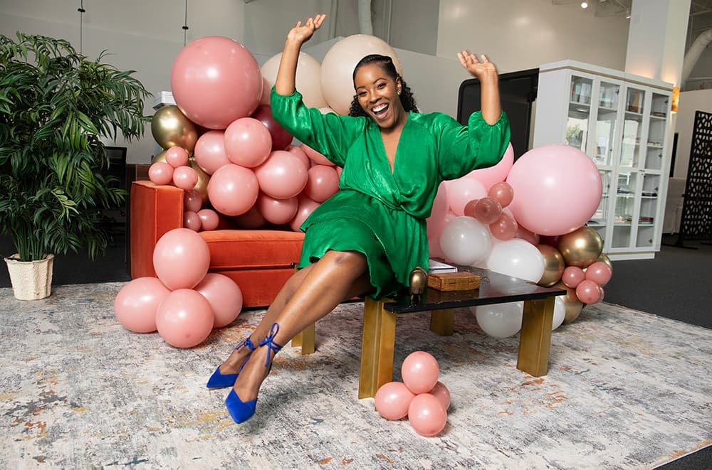 Person in a green dress sitting on a table, surrounded by pink and white balloons, with arms raised, smiling in a festive indoor setting.