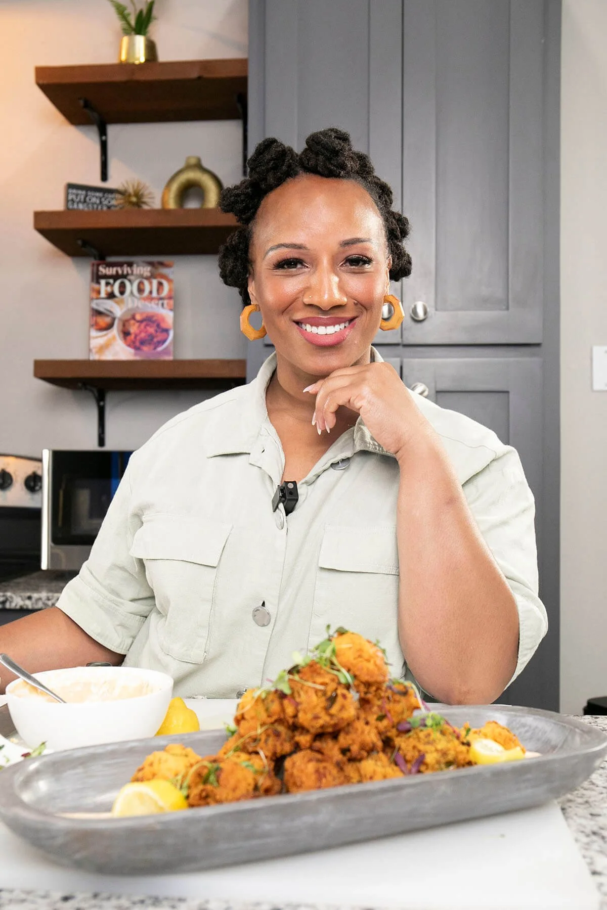 Smiling person in a kitchen, wearing a light shirt and orange earrings, posing with a tray of food, possibly fritters, garnished with lemon and herbs. Shelves and kitchen cabinets are visible in the background.