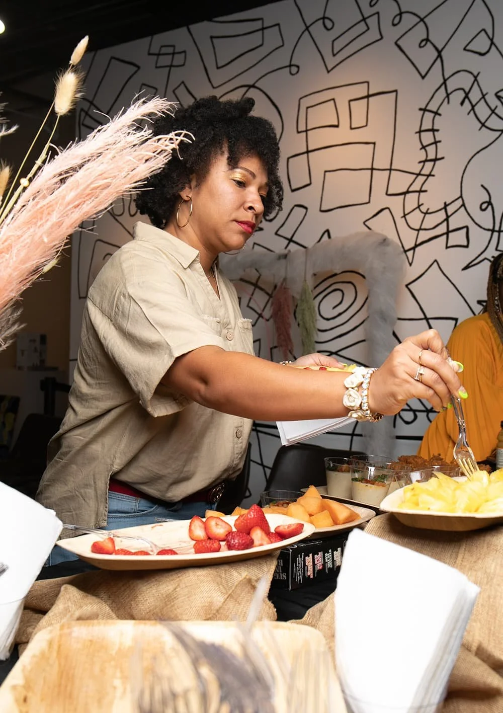 A woman in a beige shirt serves herself food from a buffet table with dishes of fruits and desserts, set against a background with abstract line drawings.