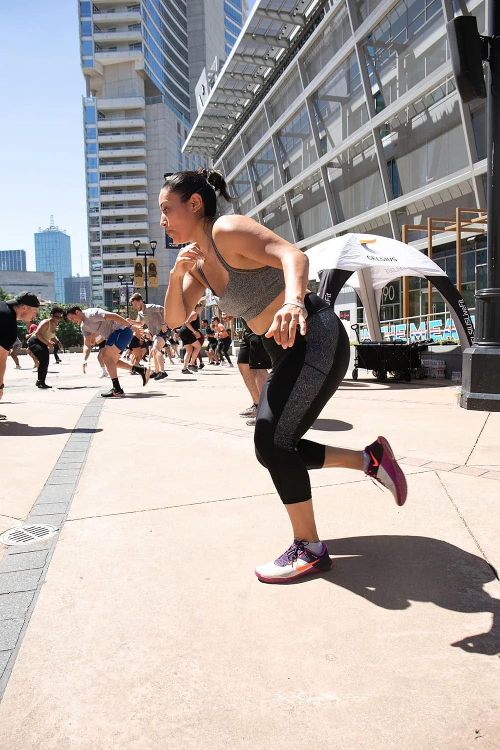 A group of people participating in an outdoor fitness class in an urban area with tall buildings in the background. A woman in athletic gear is in the foreground, focusing on her workout.