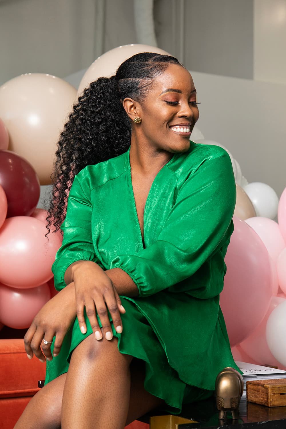 Smiling woman in a green dress sitting in front of a backdrop with pink and white balloons.