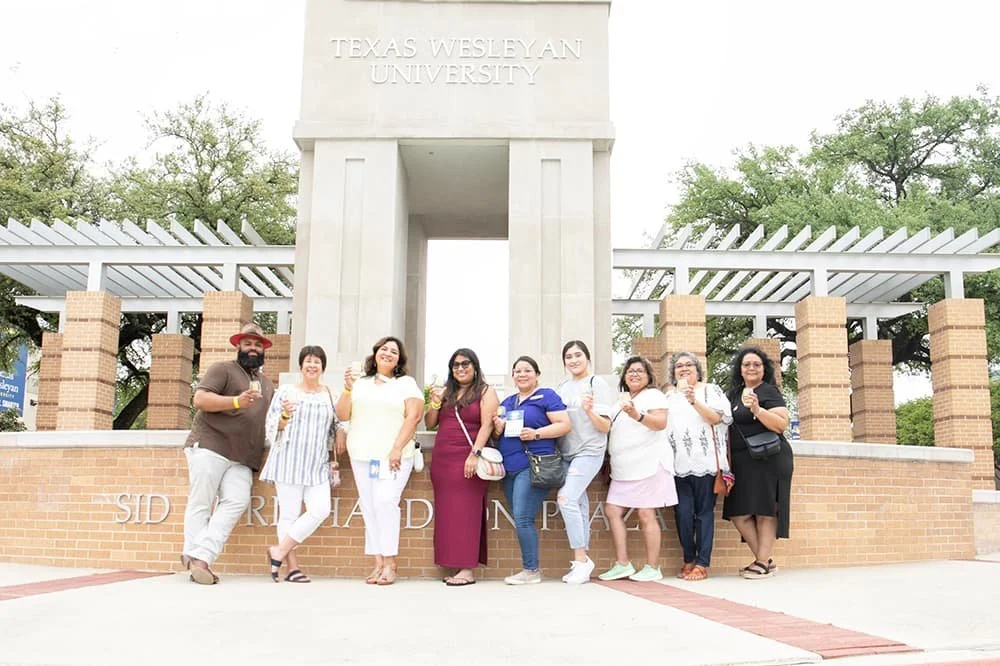 A group of people posing in front of the Texas Wesleyan University sign, standing near a brick and stone structure with a pergola, surrounded by trees.