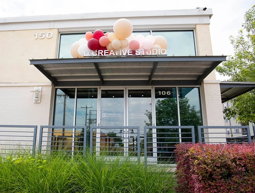 Exterior of a building with the text "Creative Studio" above the entrance, decorated with colorful balloons. The building is surrounded by greenery and has a modern design with glass windows and a metal awning.