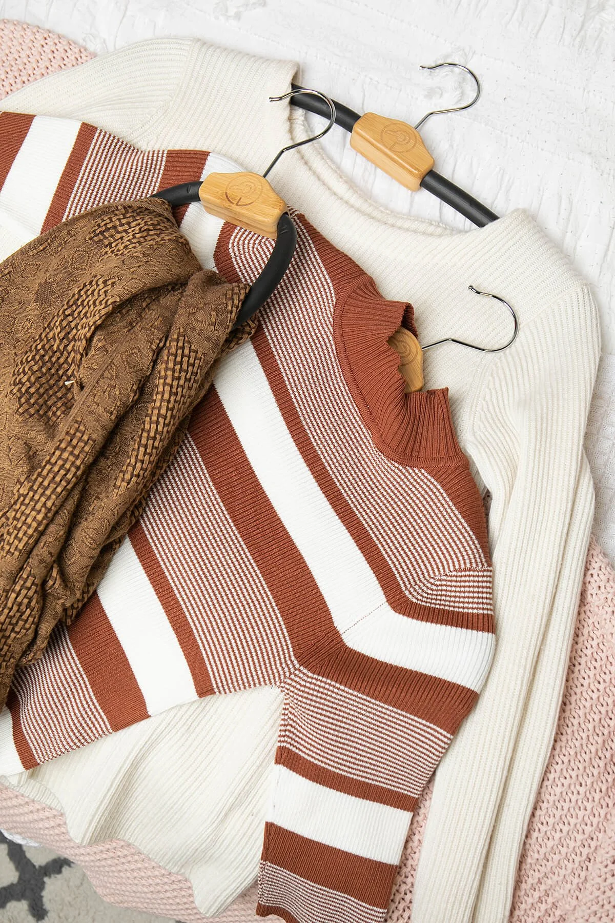 Folded sweaters on hangers, featuring white, brown, and striped designs, arranged on a textured surface.