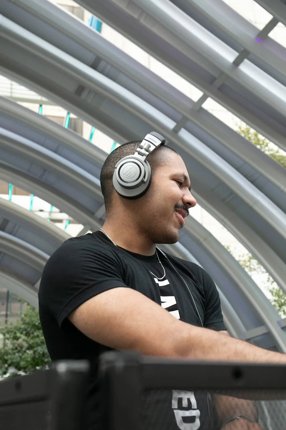 Man wearing headphones and a black t-shirt, smiling in front of a modern architectural background.