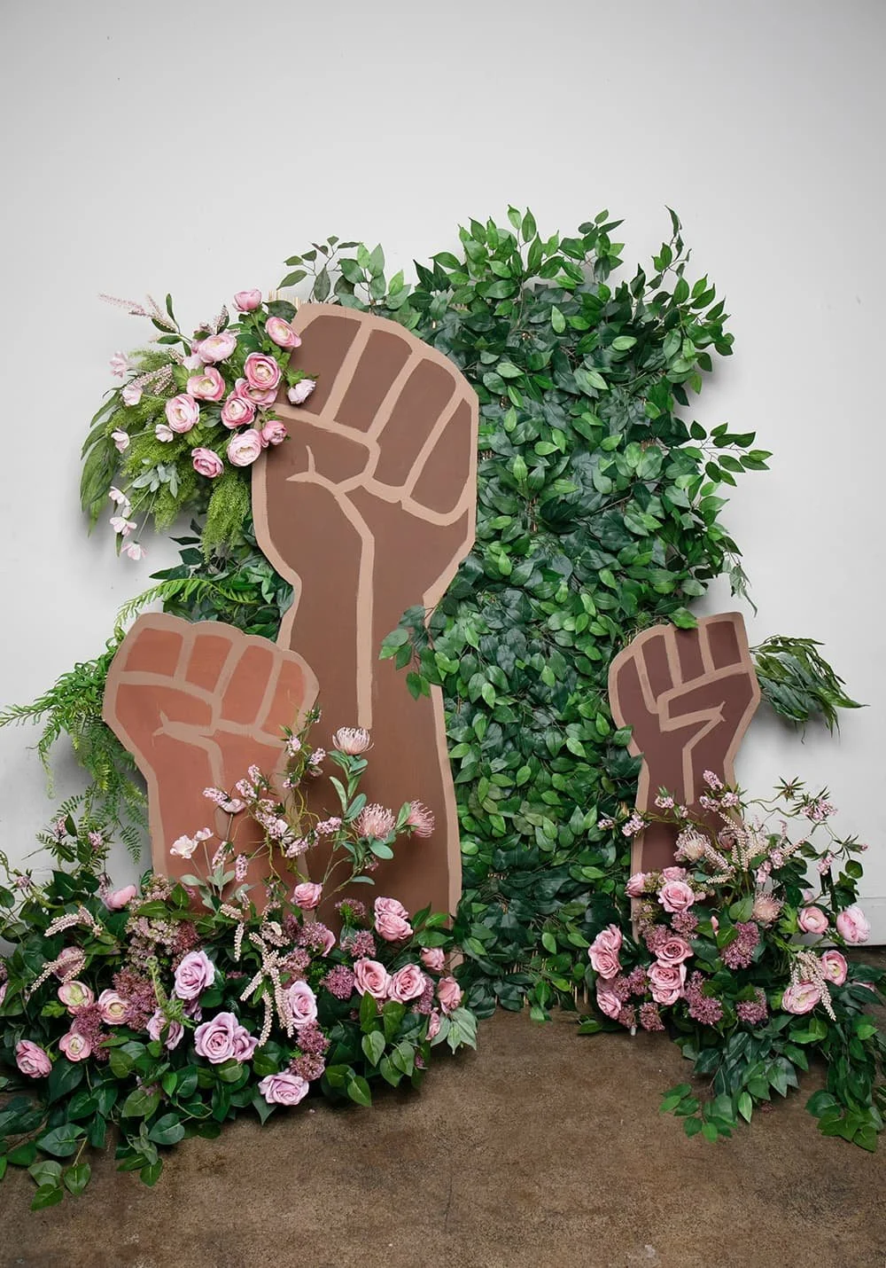 Three clenched fist cutouts surrounded by green foliage and pink flowers, symbolizing unity and strength.