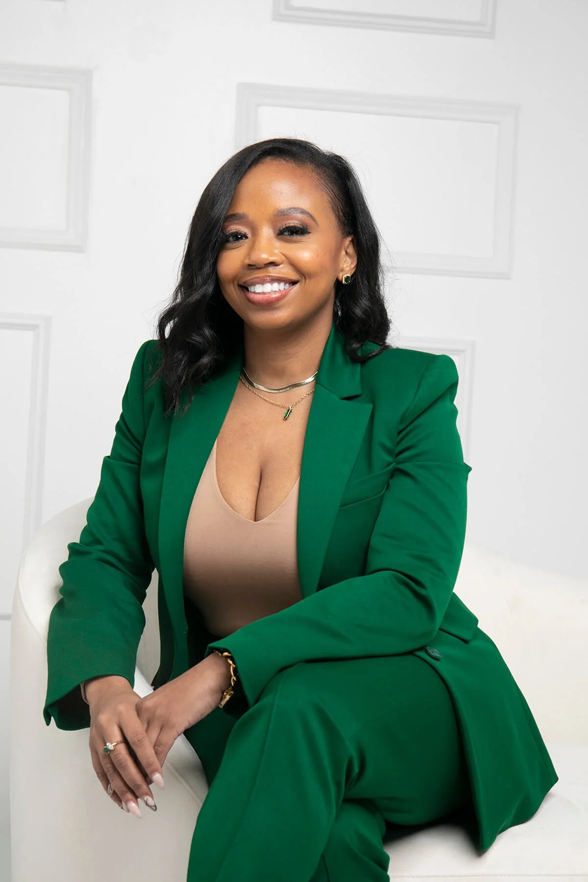 Woman wearing green suit and beige top, sitting and smiling in a light-colored room.