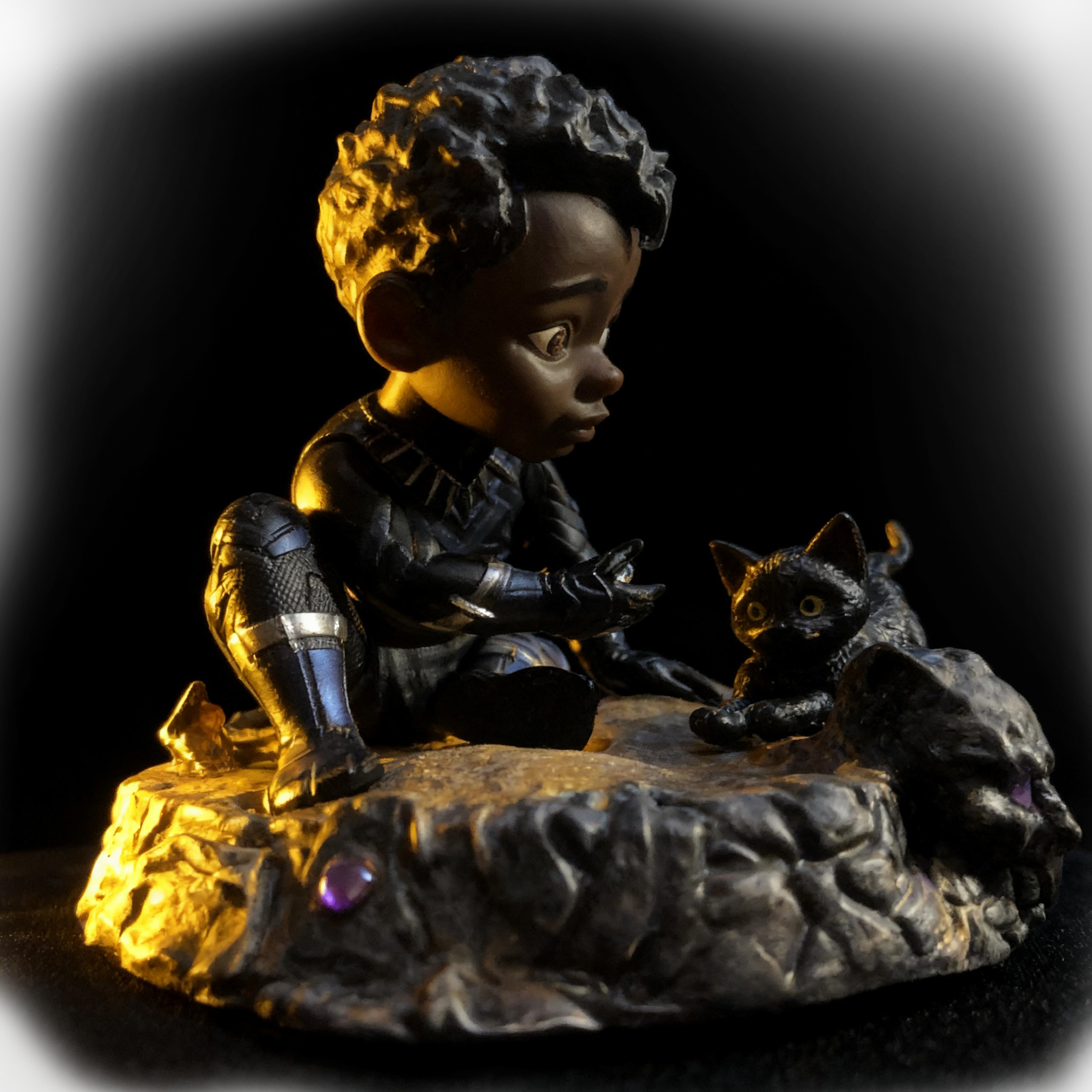 A figurine of a young Black boy with curly hair, dressed in black, sitting on a textured rocky surface, looking at a black cat with large yellow eyes, against a dark background with warm light highlighting the figures.