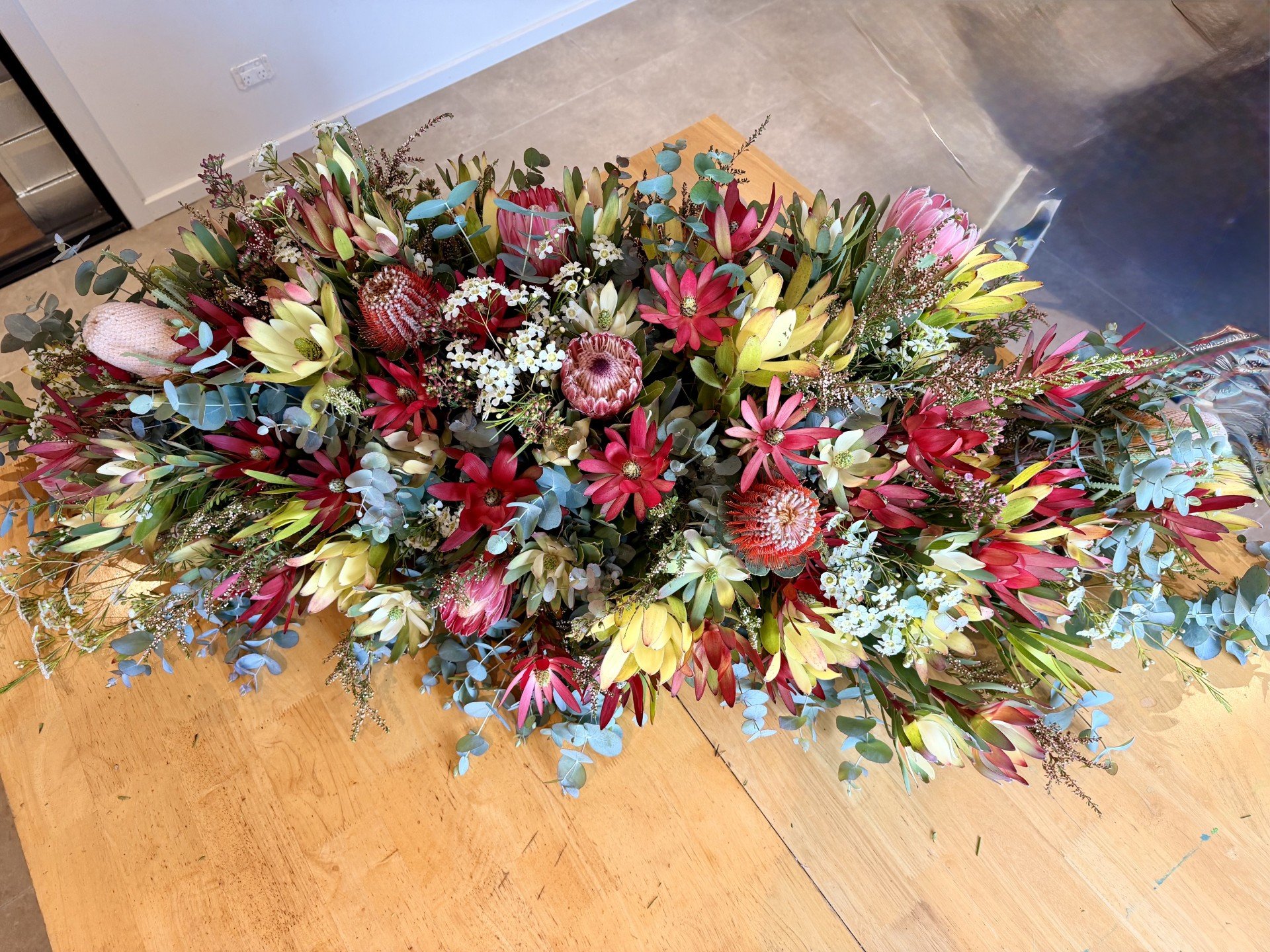 A large bouquet of assorted flowers with red, pink, white, and yellow blooms and green foliage, resting on a wooden surface.