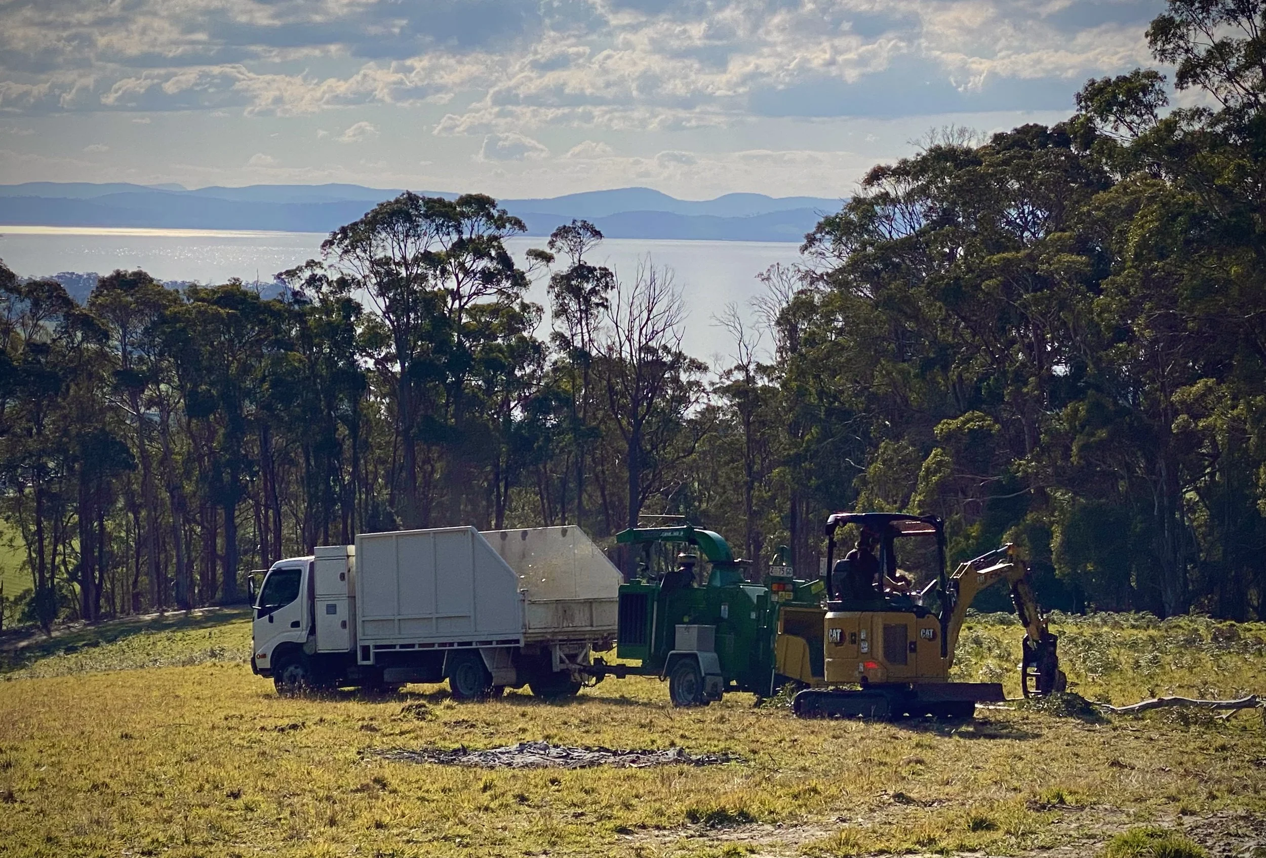 Construction equipment and vehicles in a grassy area with a backdrop of trees, water, and distant mountains under a partly cloudy sky.