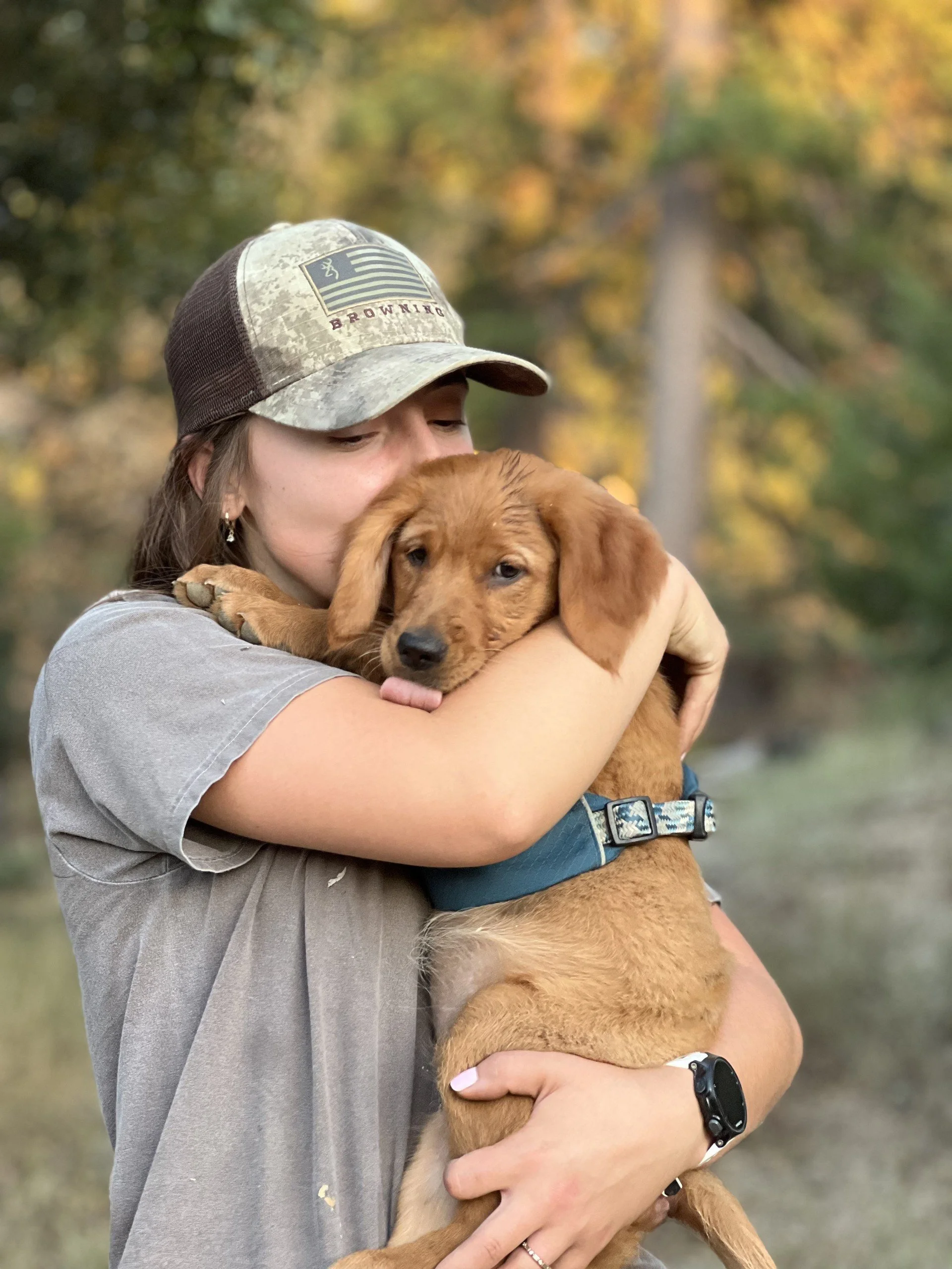A woman wearing a camouflage cap and a gray T-shirt hugging a brown puppy outdoors with trees in the background.