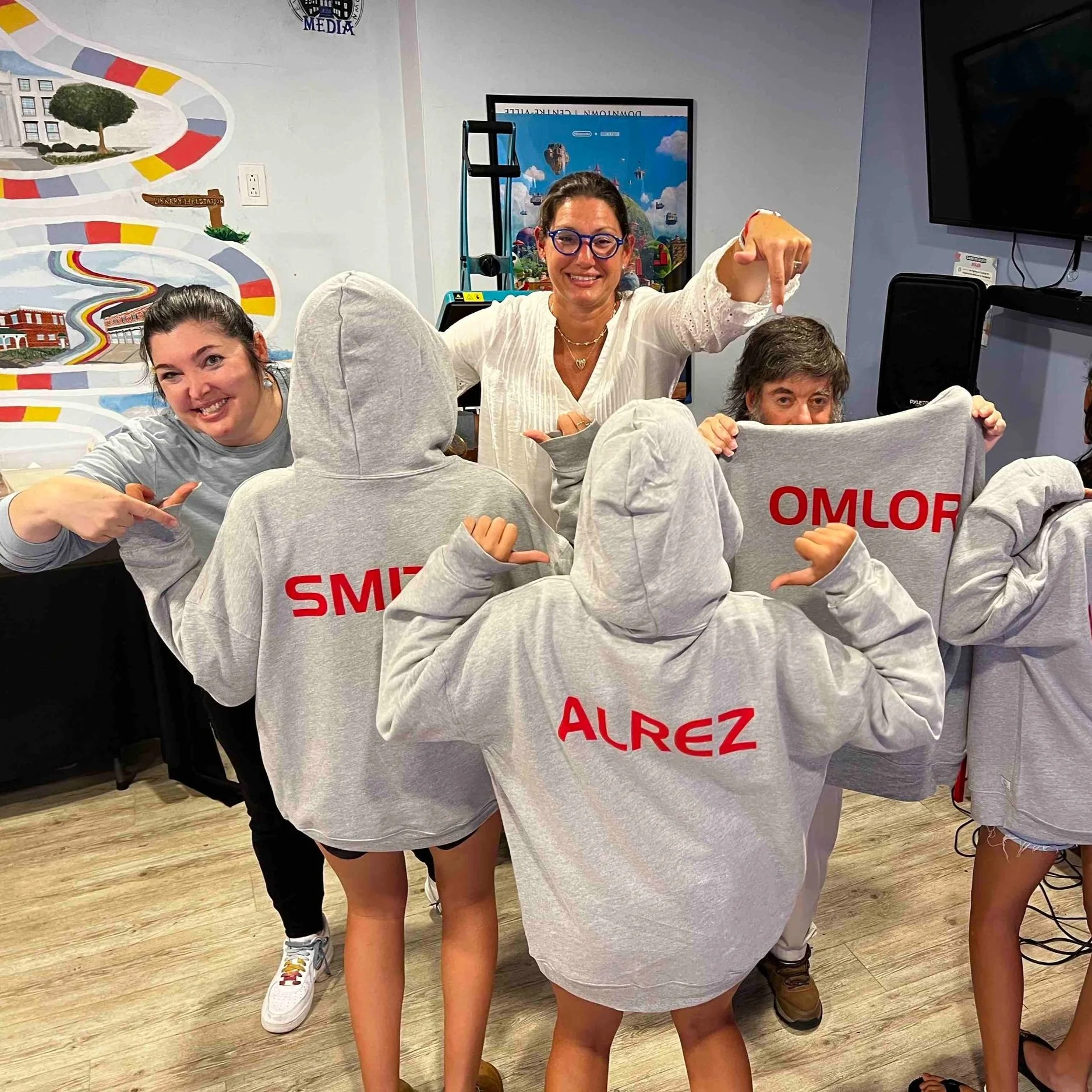 Group of people indoors, with some wearing gray hoodies with red text on the back, and smiling at the camera. One woman is pointing at the hoodies, and another is holding up the hoodies with their names printed on them.
