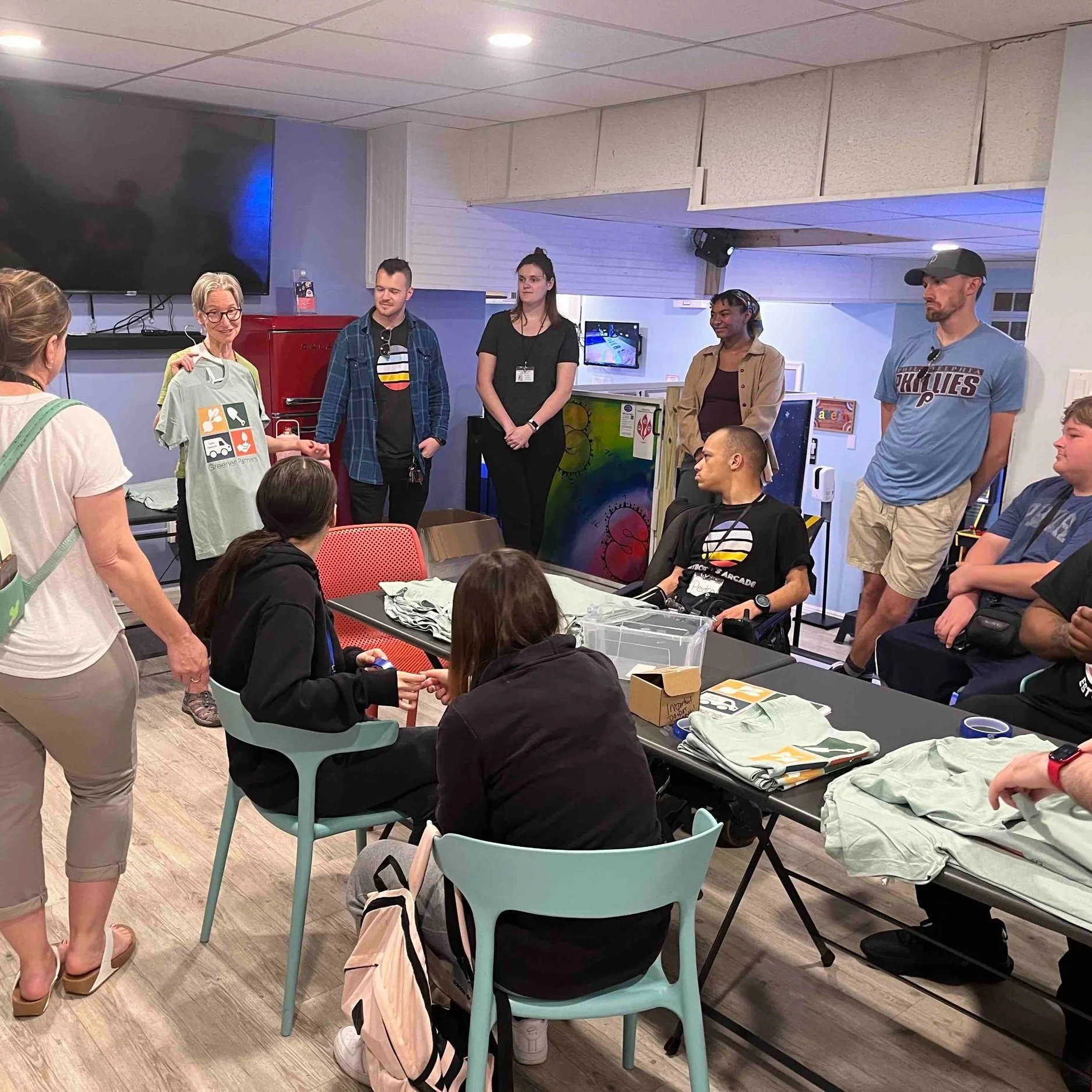 A group of people gathered in a room, with some sitting at tables and others standing, listening to an elderly woman holding up a T-shirt with colorful graphics. The room has a large TV on the wall and arcade game machines in the background. Others are seated around, some in wheelchairs, and are paying attention to the woman.