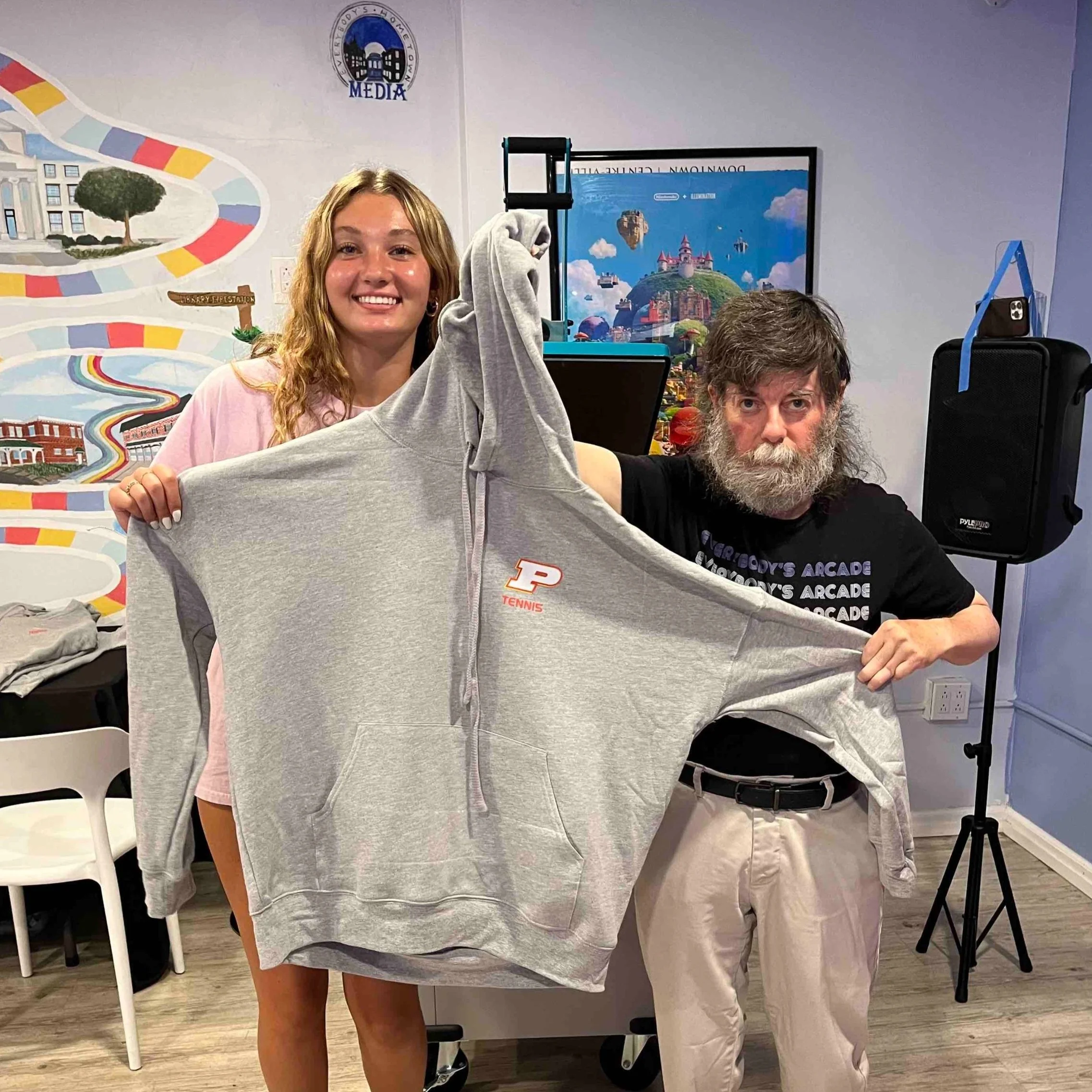 A young woman and a bearded man holding a gray Purdue University hoodie together in a room with colorful decorations and a TV screen in the background.