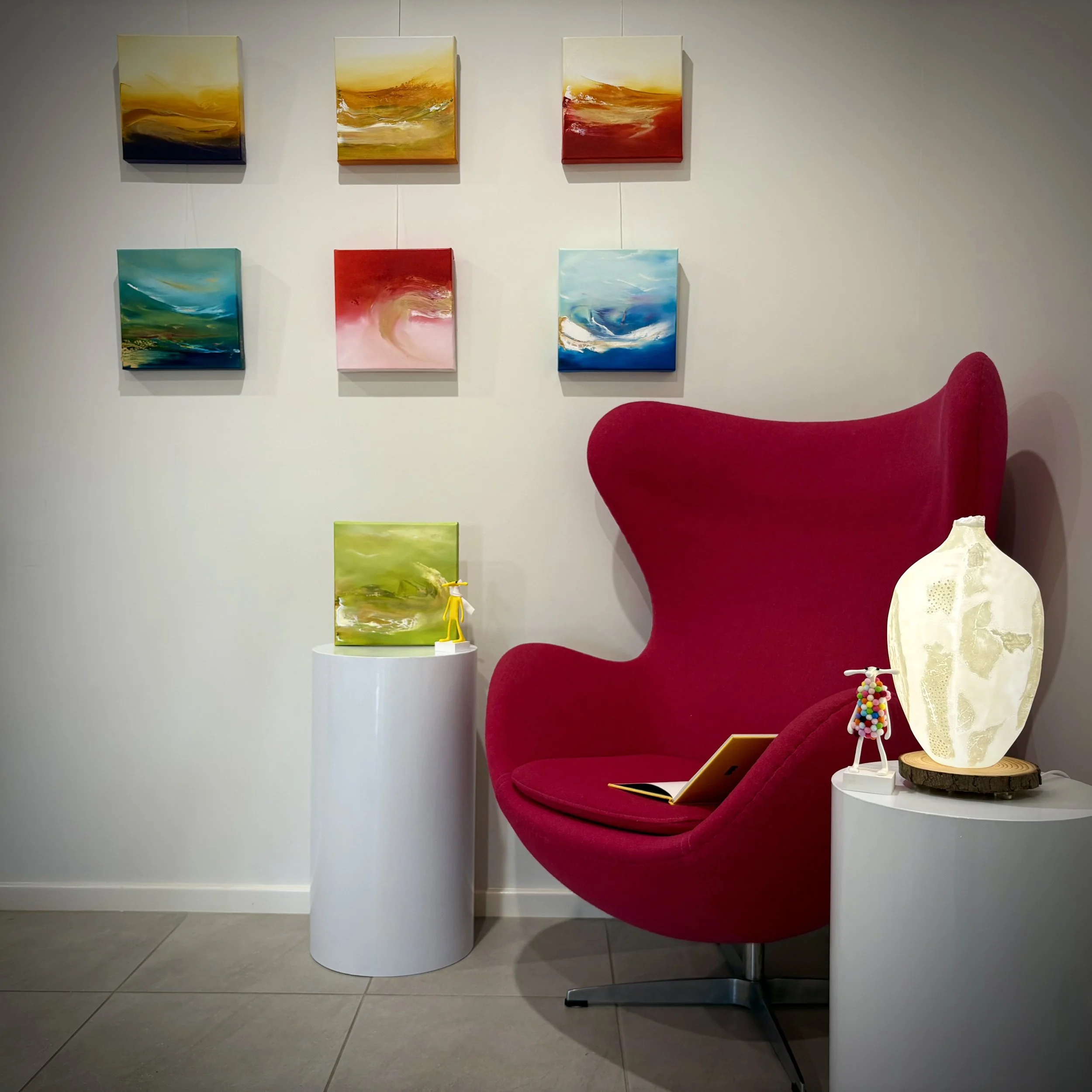 Art gallery corner with six colorful abstract paintings on the wall, a red wingback chair, two white cylindrical side tables with decorative objects, including a small yellow sculpture, a glowing white vase, and a small figurine, and an open book on the chair's seat.